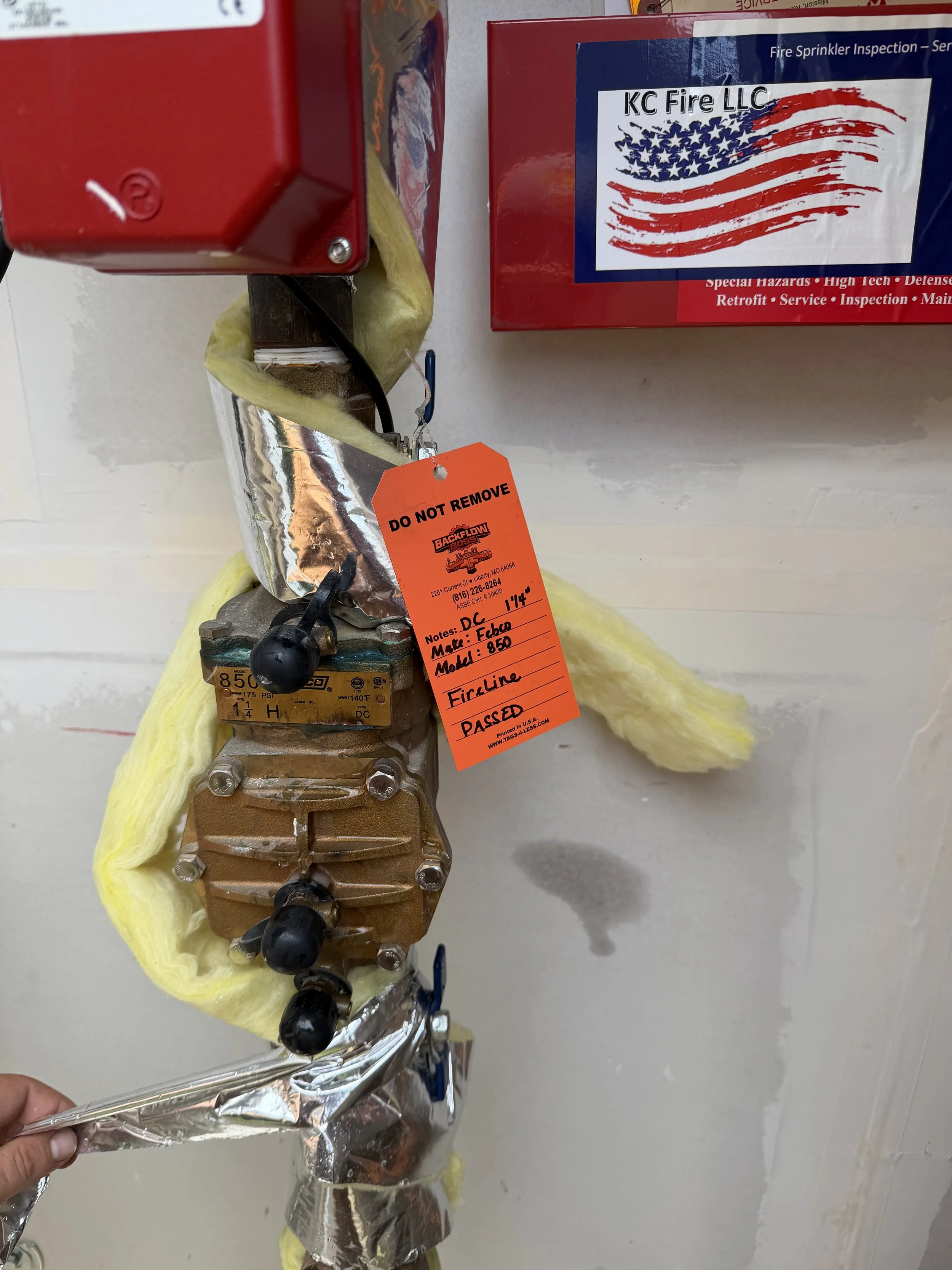 A fire sprinkler valve wrapped in yellow insulation and foil, with an orange tag attached reading 'DO NOT REMOVE' and handwritten notes indicating it has passed a fire line test, located on a white wall near a red fire alarm box and a red box with a sticker of the American flag.