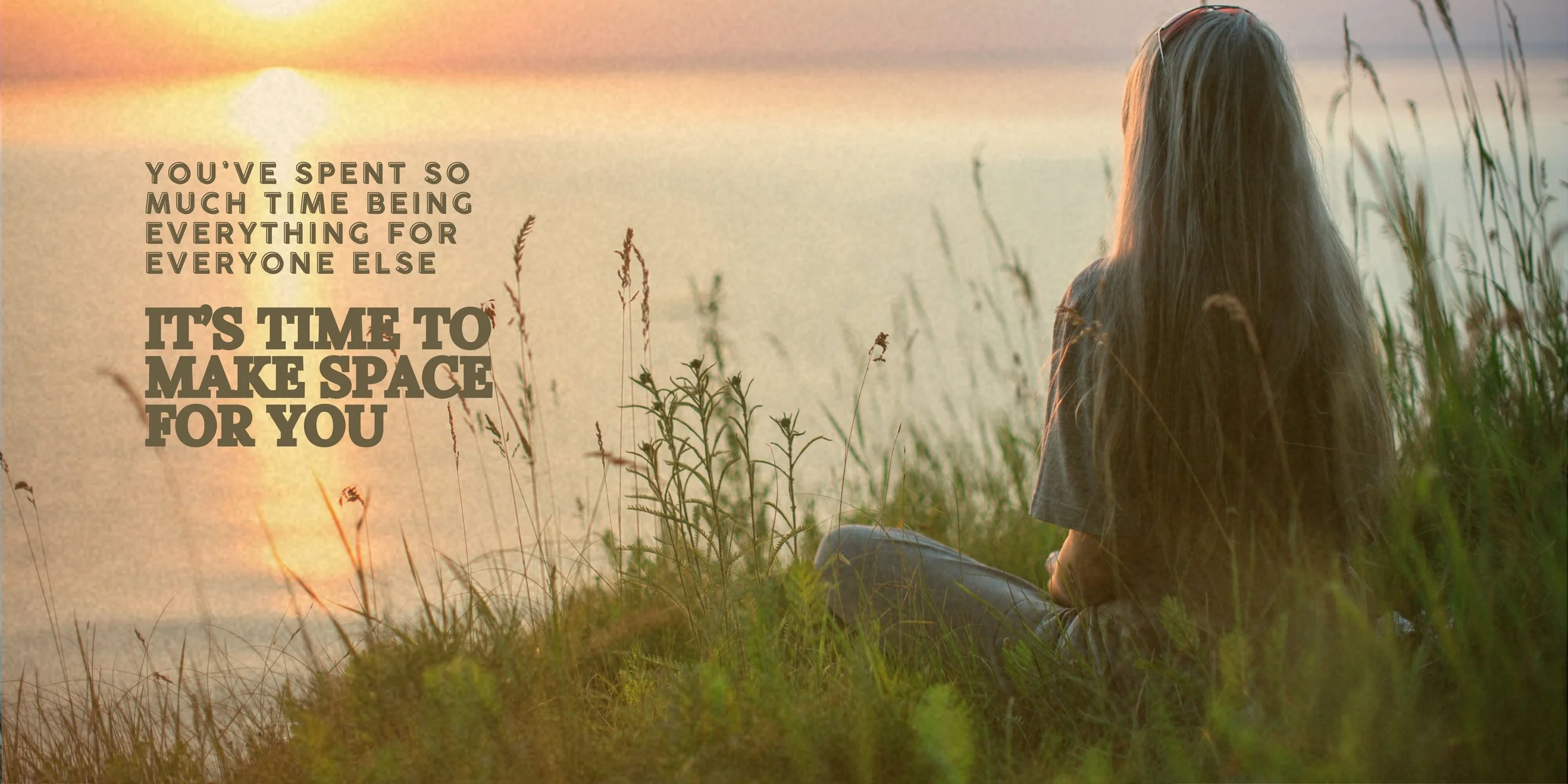 A woman with long hair, sitting on grass near a body of water, watching a sunset with her back to the camera.