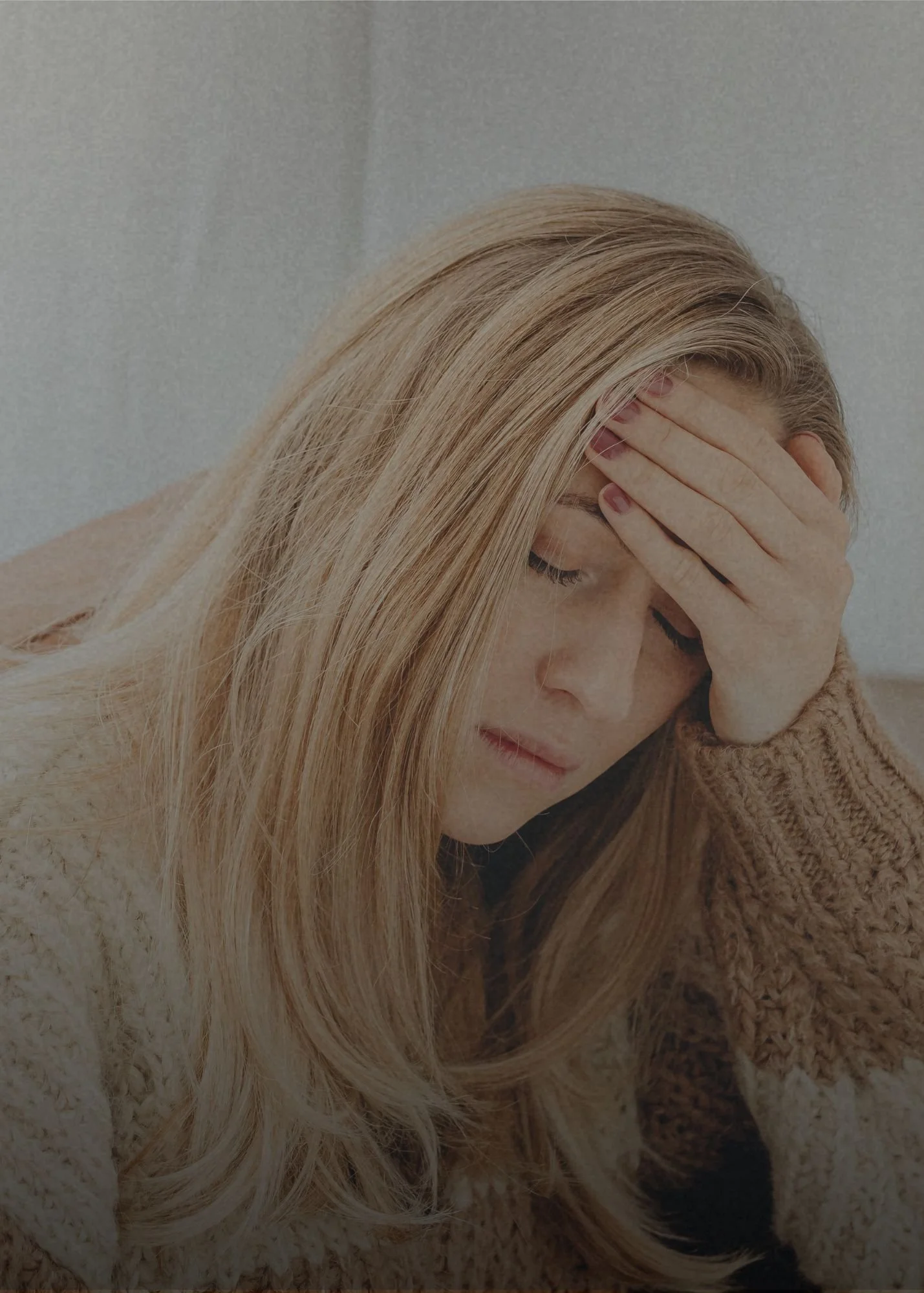 A woman with long blonde hair, wearing a beige sweater, holding her head with her hand and appearing distressed.