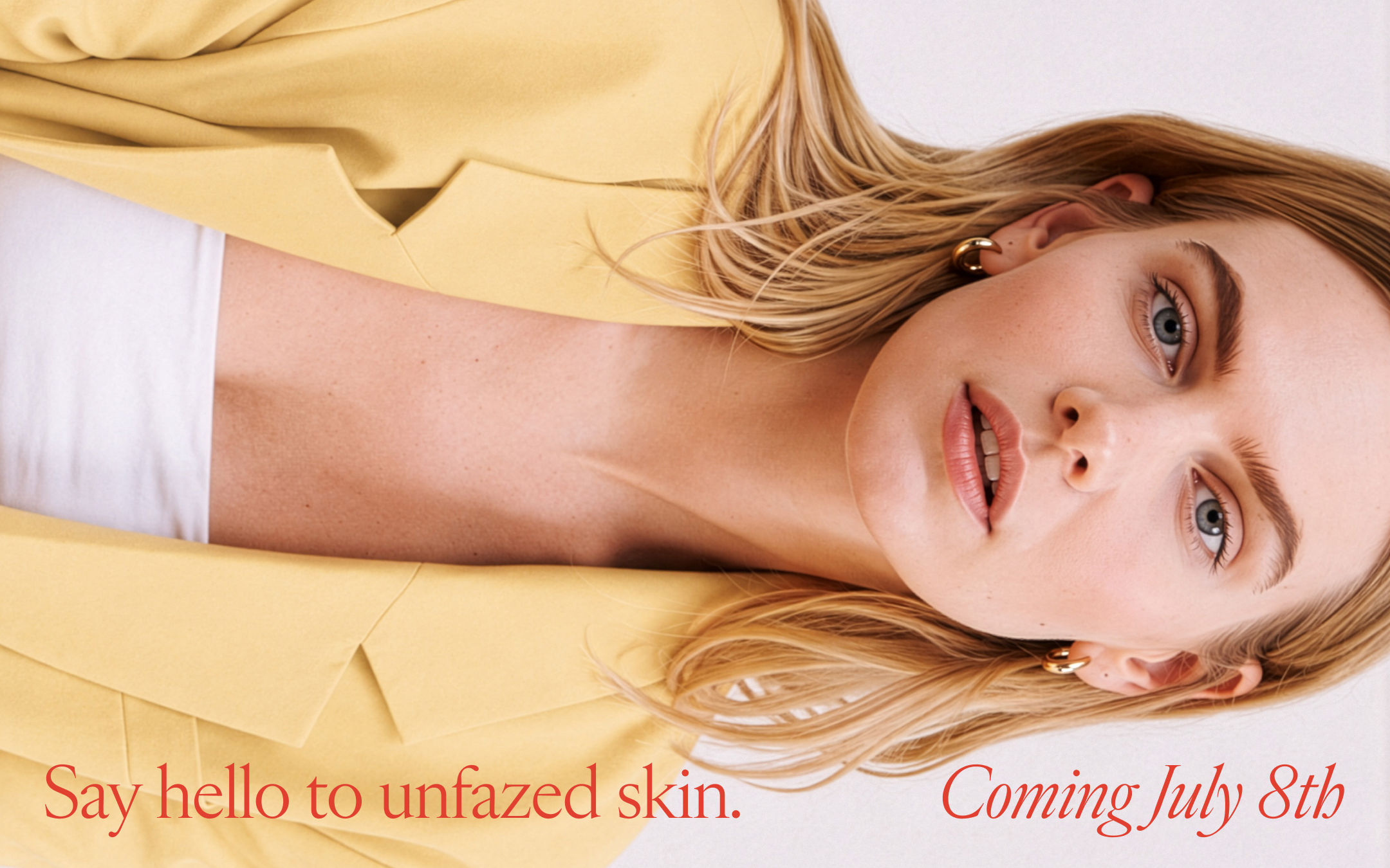 Close-up of a woman with blonde hair, wearing a yellow blazer over a white shirt, and gold hoop earrings, showcasing her unfaszed skin.