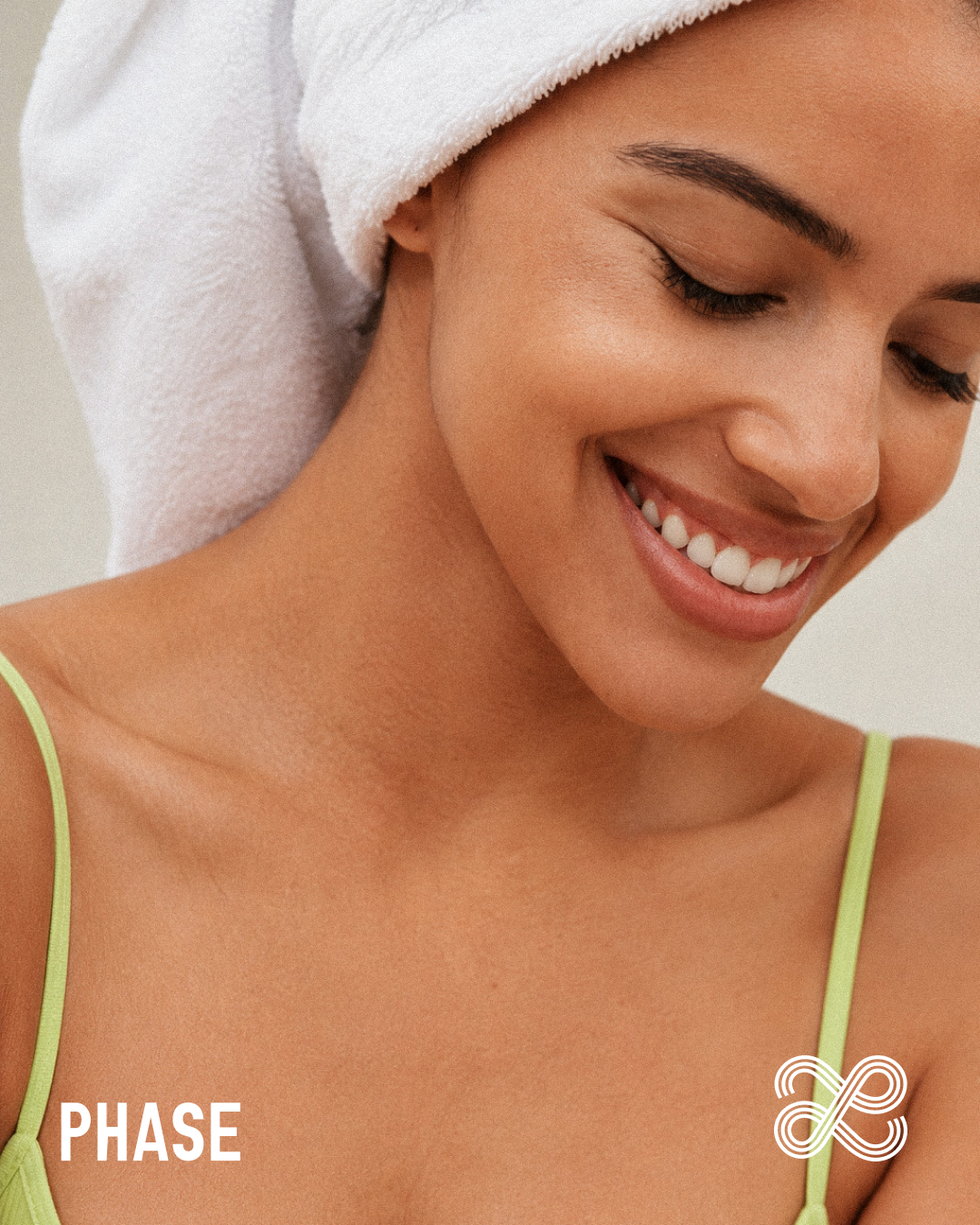 A woman with a towel wrapped around her head, smiling with closed eyes and showing her teeth with the Phase skincare branding in each of the bottom corners.
