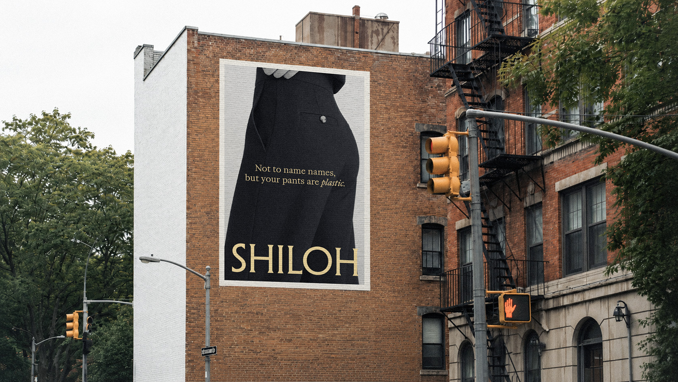 A large billboard on a brick building shows a black and white image of a person's lower body wearing black pants with a quote reads, 'Not to name names, but your pants are plastic.' The building has fire escape stairs on its side, and a traffic light and street signs are visible below. Trees are in the background.