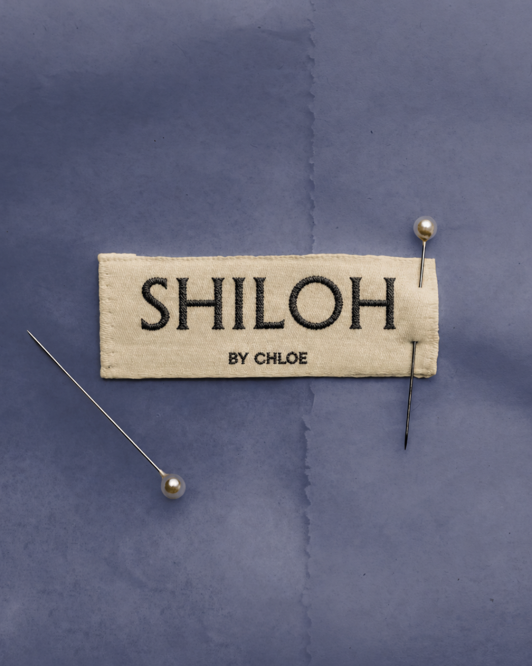 Garment label with the 'SHILOH BY CHLOE' logo embroidered in black on cream fabric, pinned to a blue tissue paper background with two straight pins.