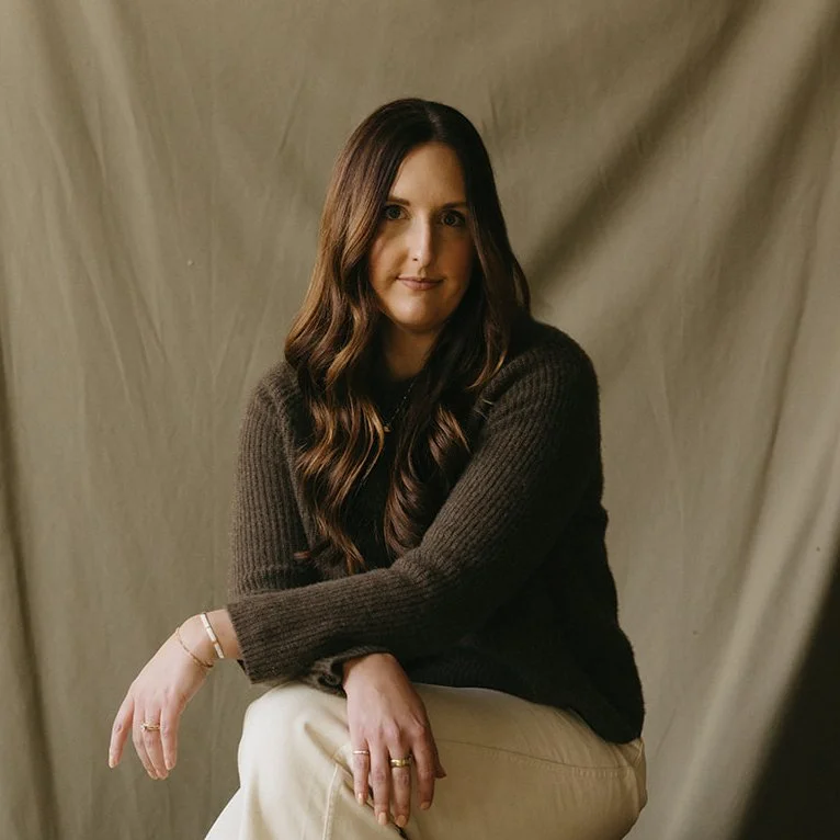 A woman with long wavy brown hair sitting against a brown fabric backdrop, wearing a dark brown sweater and light-colored pants, with relaxed posture and subtle smile.
