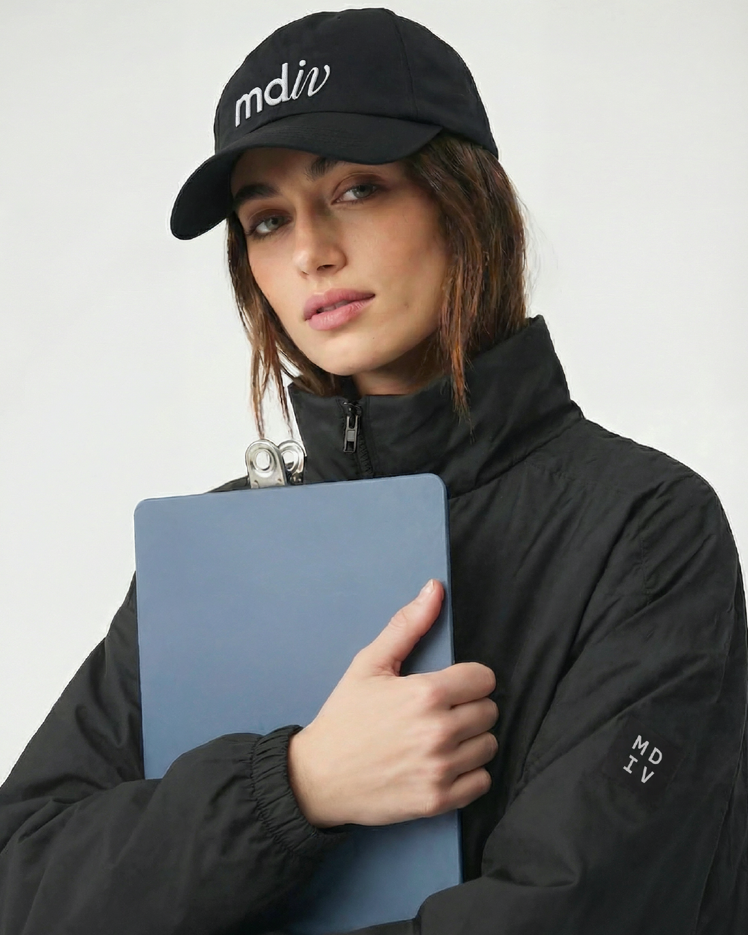 Young woman with short brown hair wearing a branded black baseball cap and black jacket holding a blue clipboard, posing against a plain white wall.