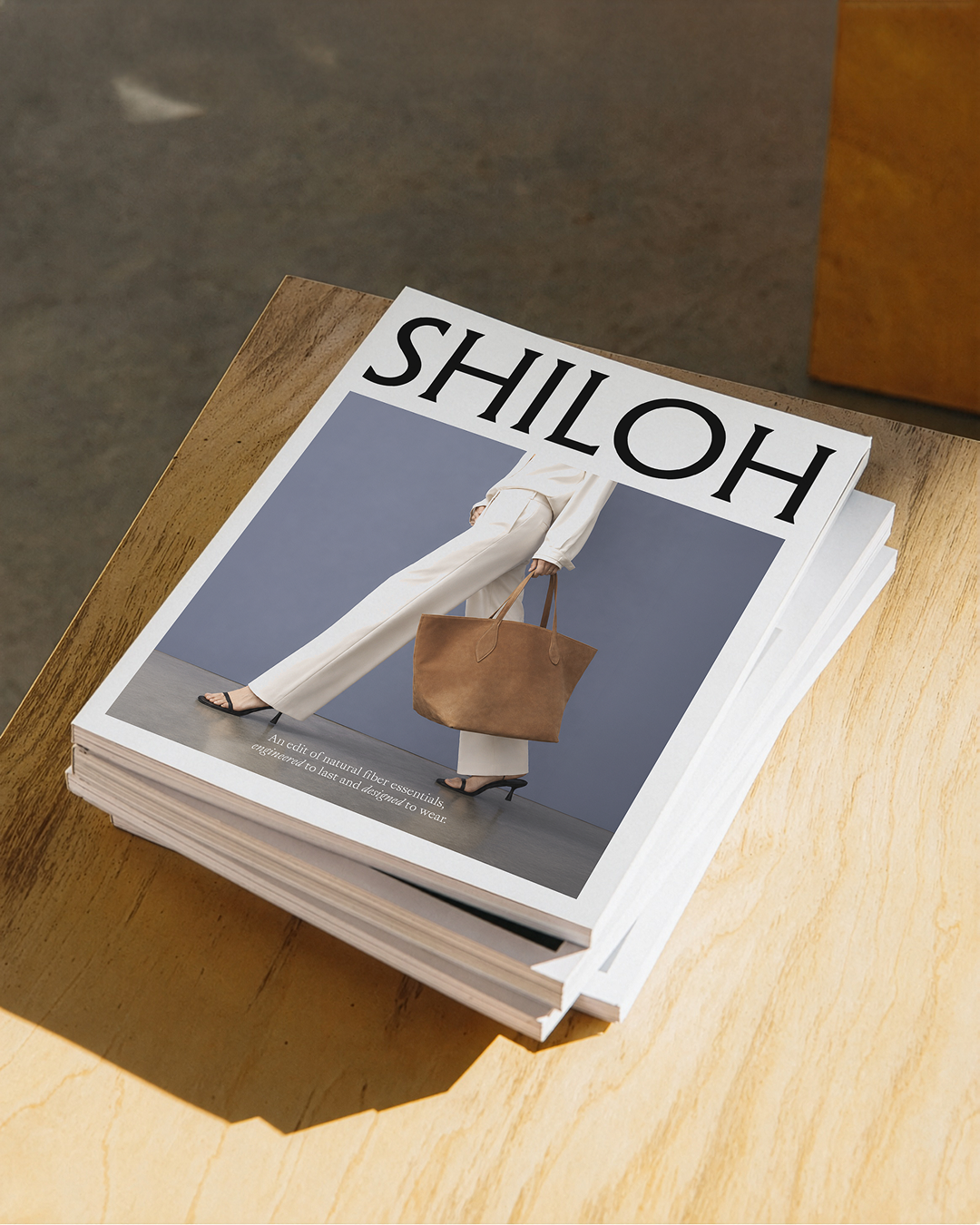 Stack of branded lookbooks for Shiloh by Chloe fashion brand, featuring a model in white pants holding a large brown bag in front of a blue background.