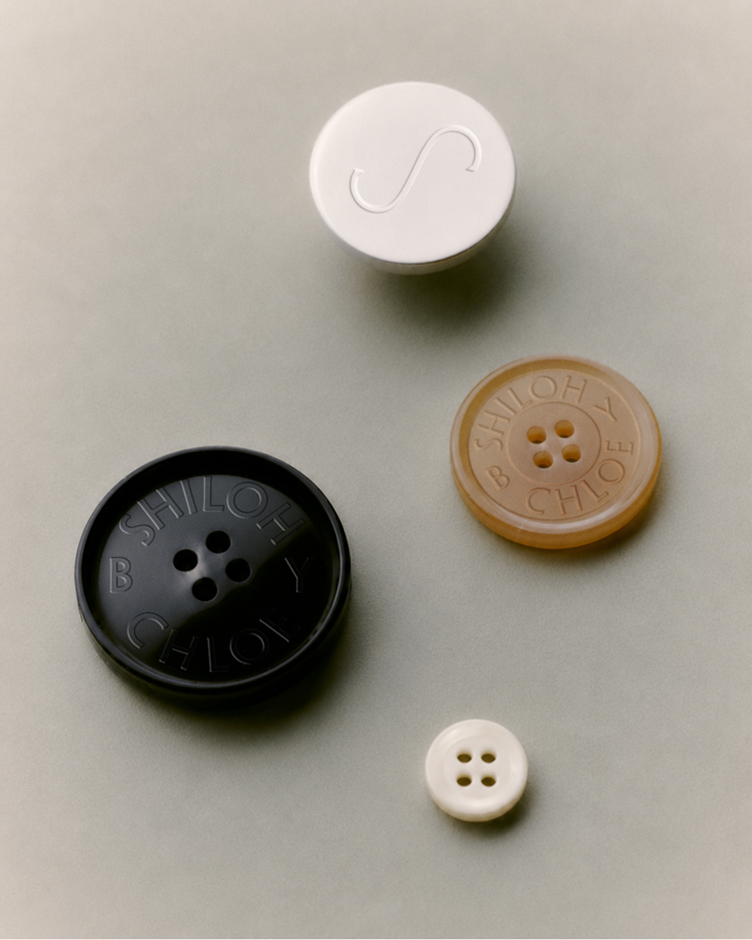 Four different colored buttons, including a silver, a tan, a black, and a small white button, are arranged on a light-colored surface, each with an imprinted fashion brand's logo on top.