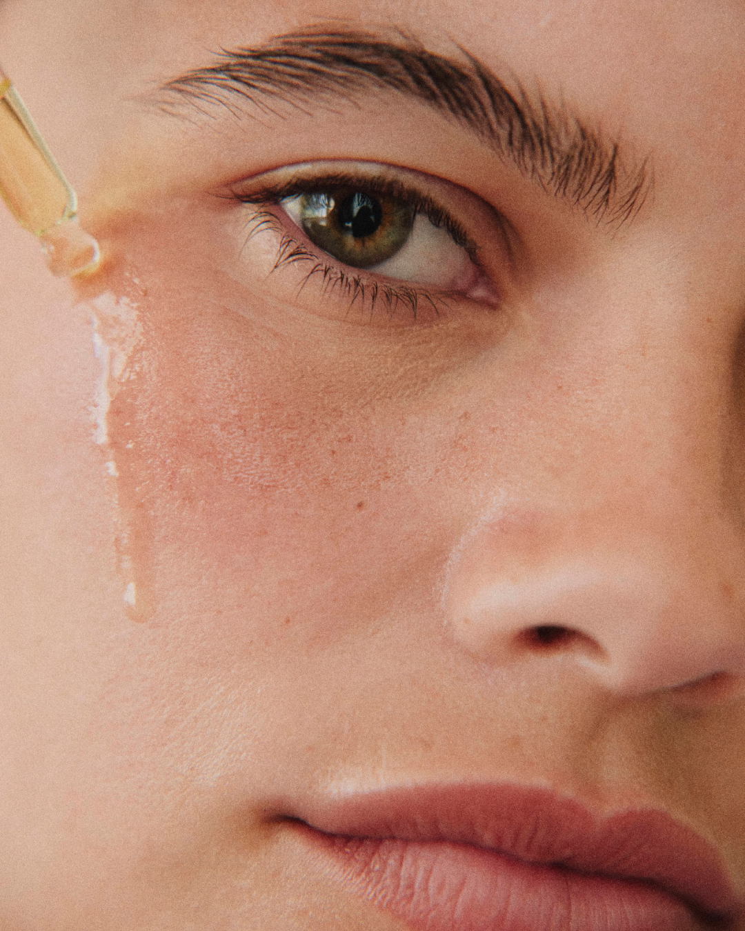 Close-up of a woman's face with a dropper applying a serum or liquid to her cheek.
