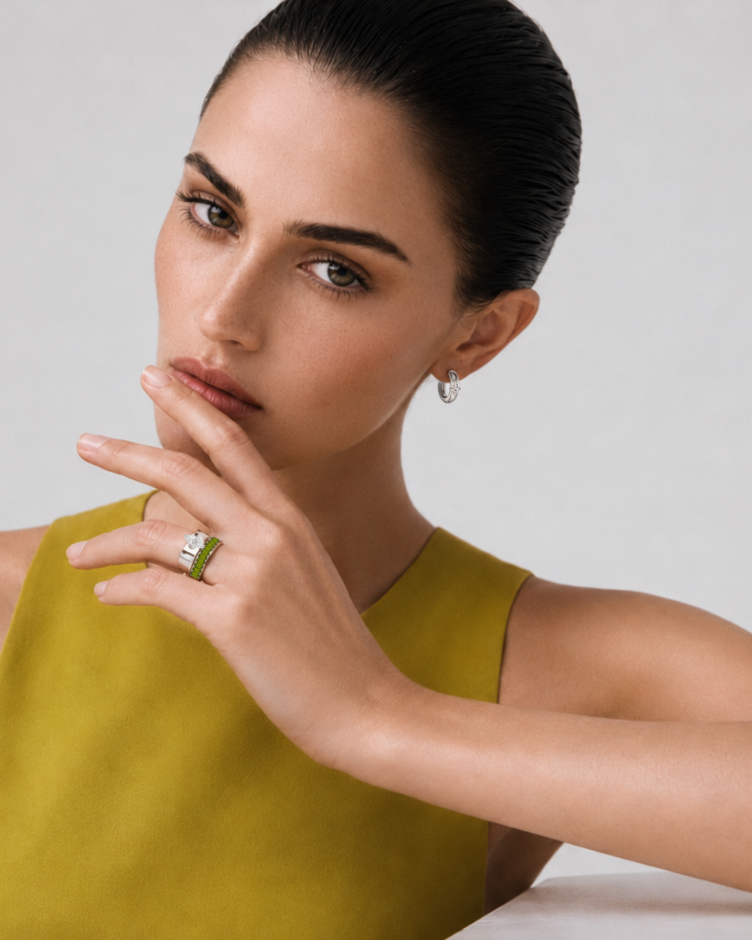 A young woman with dark, slicked-back hair wearing a chartreuse top and silver jewelry, posing with her hand near her face against a plain background.
