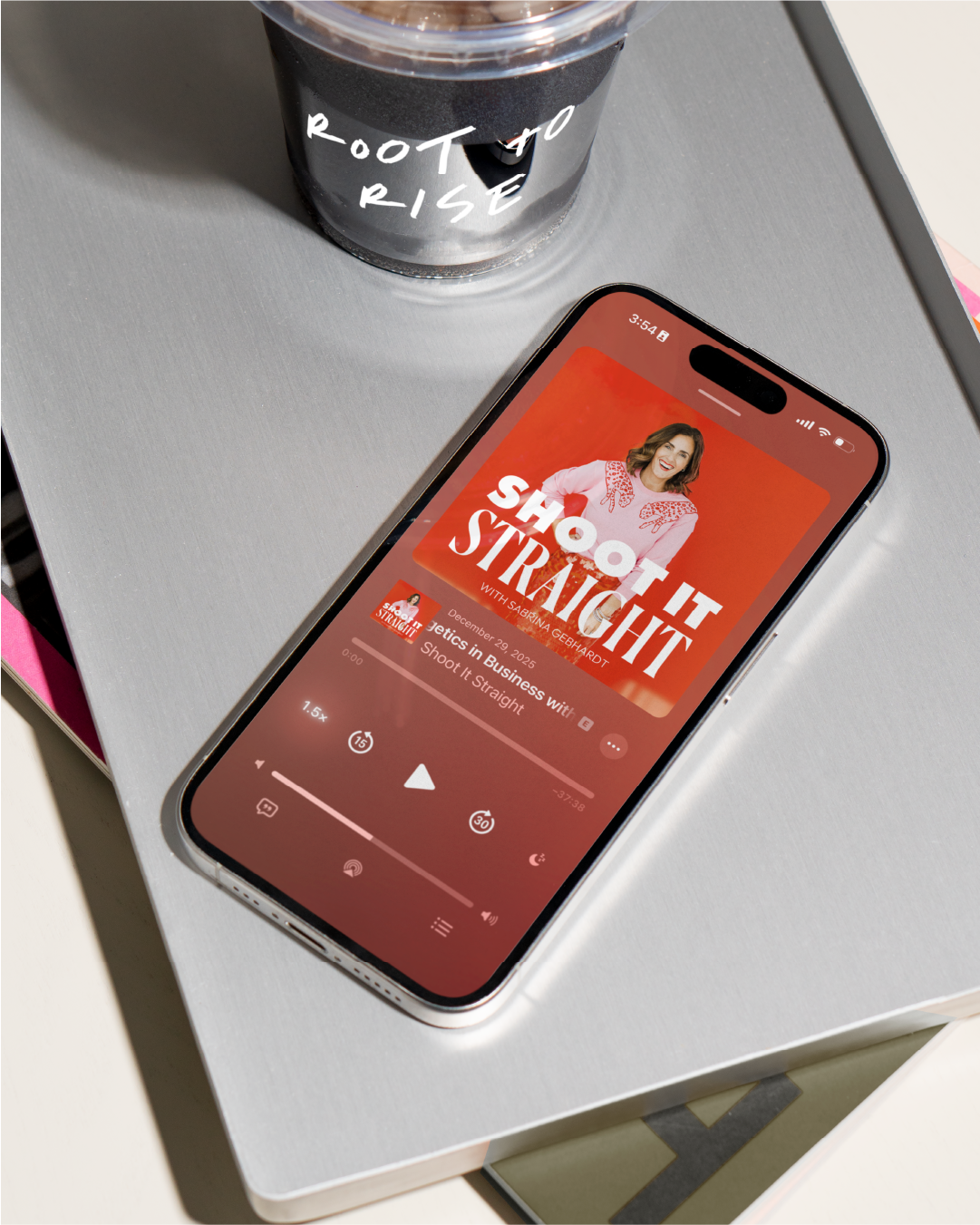 Smartphone featuring podcast cover art design titled 'Shoot It Straight with Sabrina Gebhardt' on a silver laptop, with a plastic cup labeled 'Root to Rise' nearby.