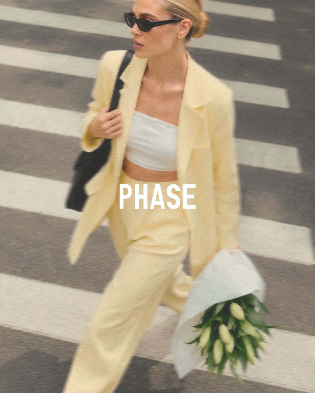 A woman wearing a yellow suit and sunglasses walking across a crosswalk while holding a bouquet of white flowers.