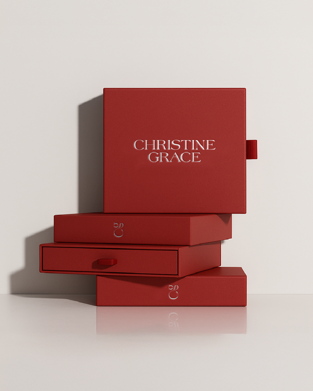 Stack of red branded packaging with silver foil printing of the Christine Grace logo, on a plain white background.