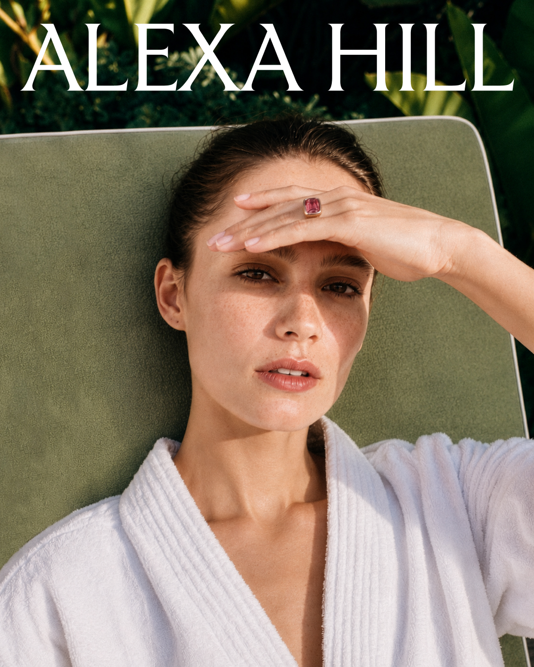 A woman lying on a green lounge chair outdoors, wearing a white robe, with her left hand on her forehead and a large pink gemstone ring on her middle finger. She has dark hair and is looking at the camera. The Alexa Hill logo is overlayed on top.