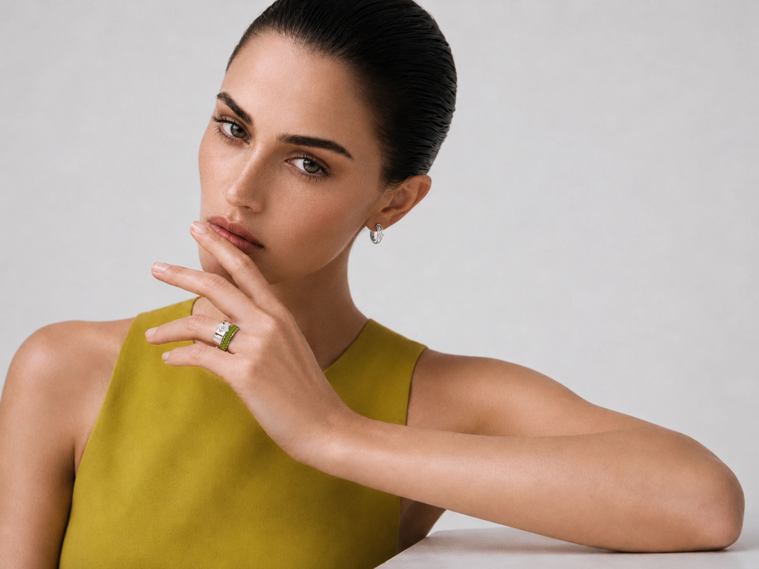 Close-up of a woman with dark hair slicked back, wearing a chartreuse top, silver earrings, and rings, against a neutral background.