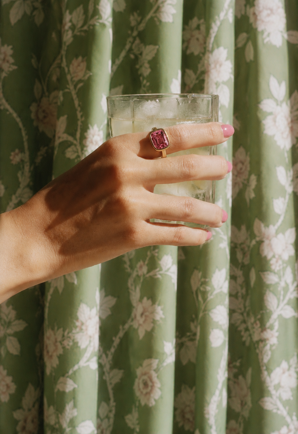 A woman's hand with pink nail polish holding a glass of a light-colored beverage with ice, wearing a large pink gemstone ring, in front of a green floral curtain.