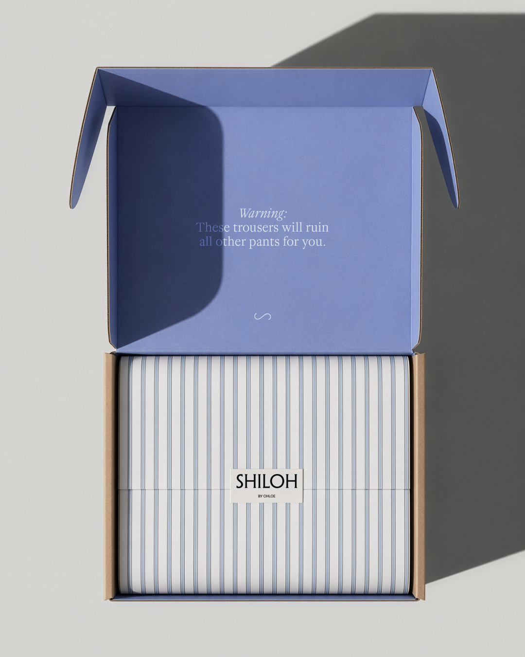 Open box containing neatly folded striped blue and white tissue paper from Shiloh by Chloe with humorous warning inside the lid about trousers ruining other pants.