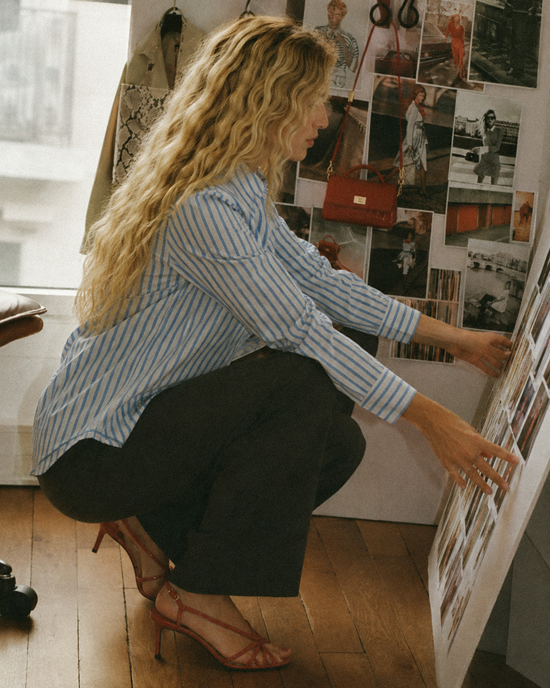 A woman with long curly blonde hair, wearing a blue and white striped shirt, black pants, and red high heels, is crouching and looking at a creative direction for a fashion shoot on a white wall.