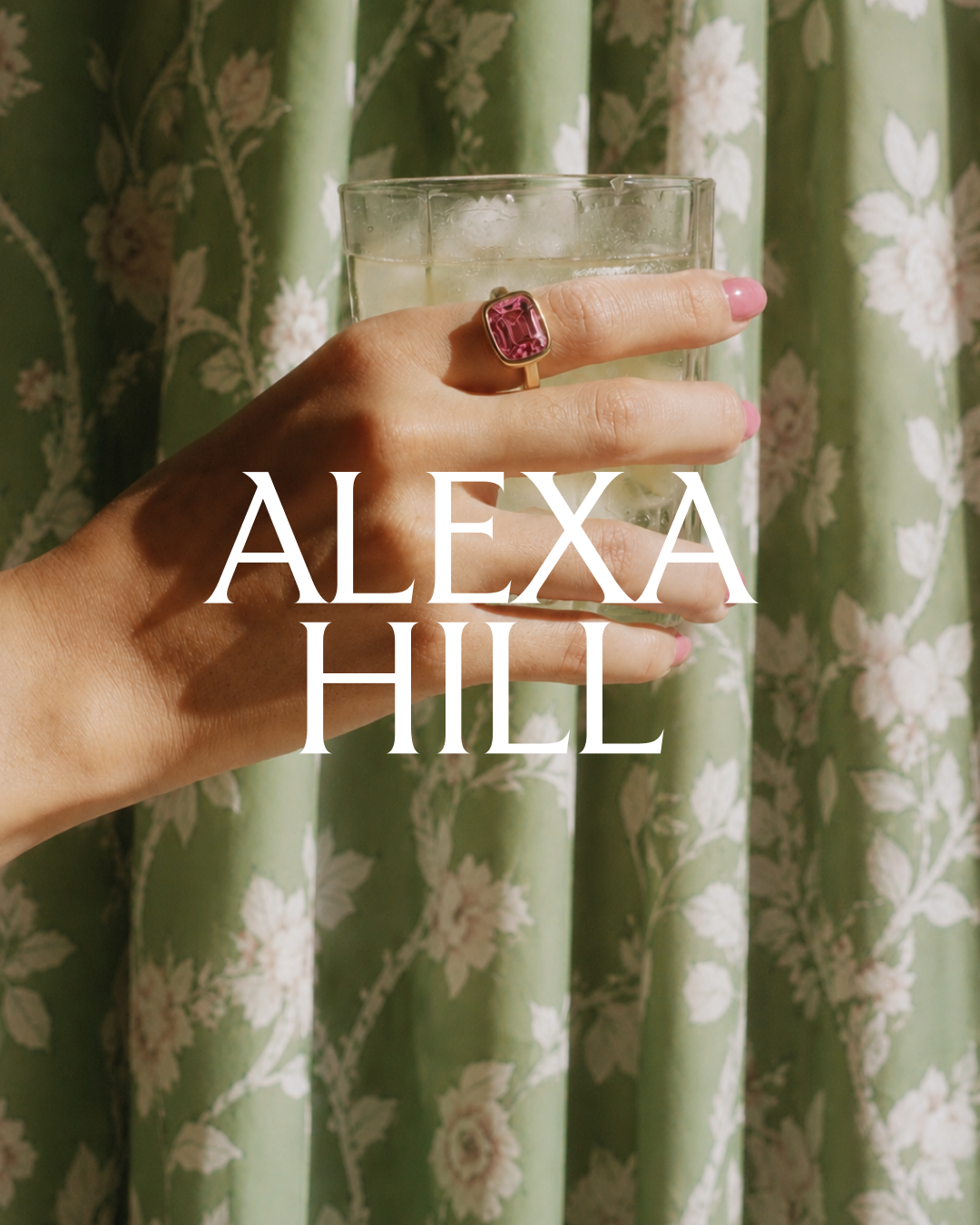 A woman's hand with pink nail polish and a pink gemstone ring, holding a glass of ice water against a green floral curtain backdrop, with the 'ALEXA HILL' logo overlayed.