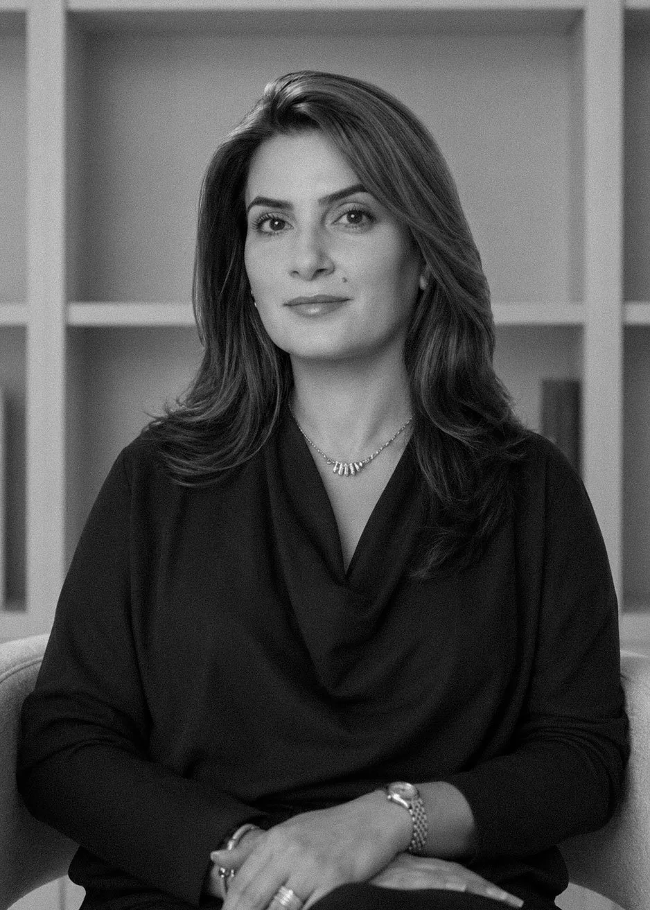 Black and white portrait of a woman with shoulder-length hair, wearing a dark top, a necklace, a watch, and rings, sitting in front of a bookshelf.