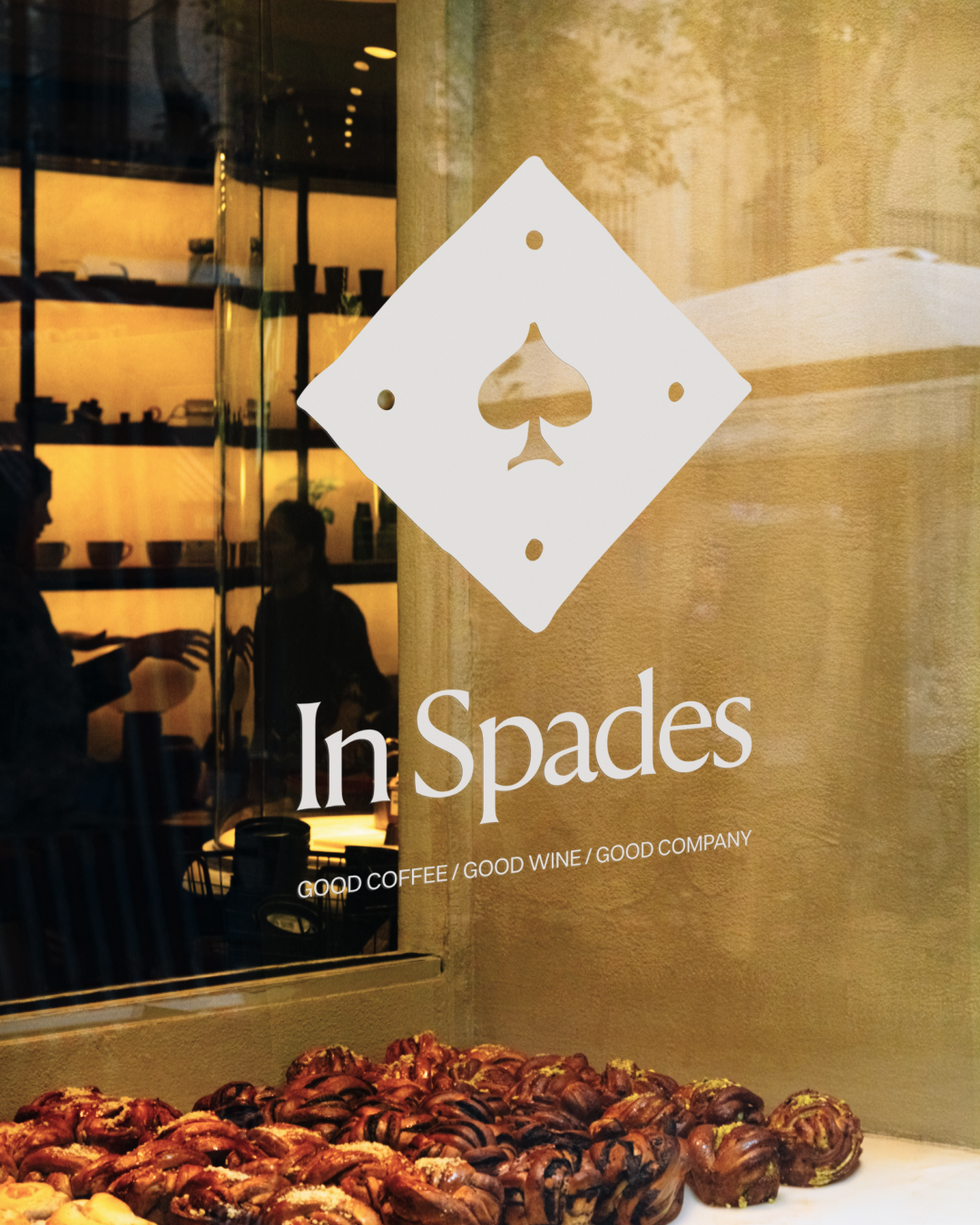 Bakery display with assorted pastries in front of a window with a sign for In Spades, a café or restaurant named In Spades that offers good coffee, wine, and company.