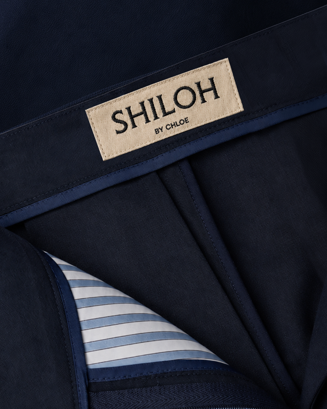 Close-up of a dark navy blue trousers label sewn onto the garment, reading "SHILOH BY CHLOE," with part of a striped lining visible.