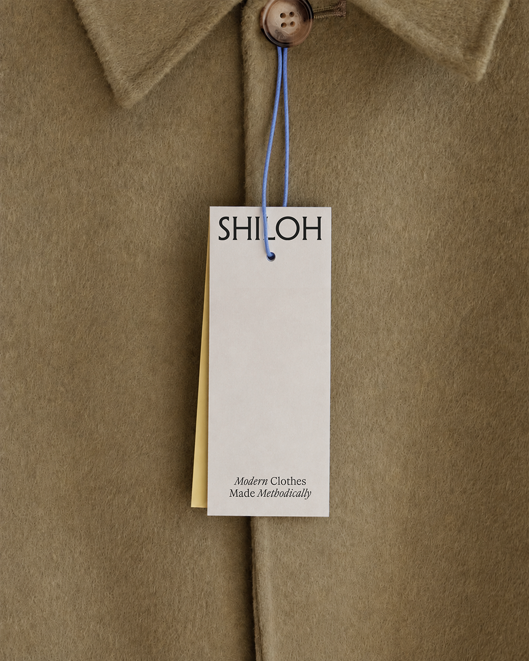 Close-up of a beige coat with a white tag hanging from a blue string with the Shiloh logo on top and the text 'Modern Clothes Made Methodically' at the bottom.