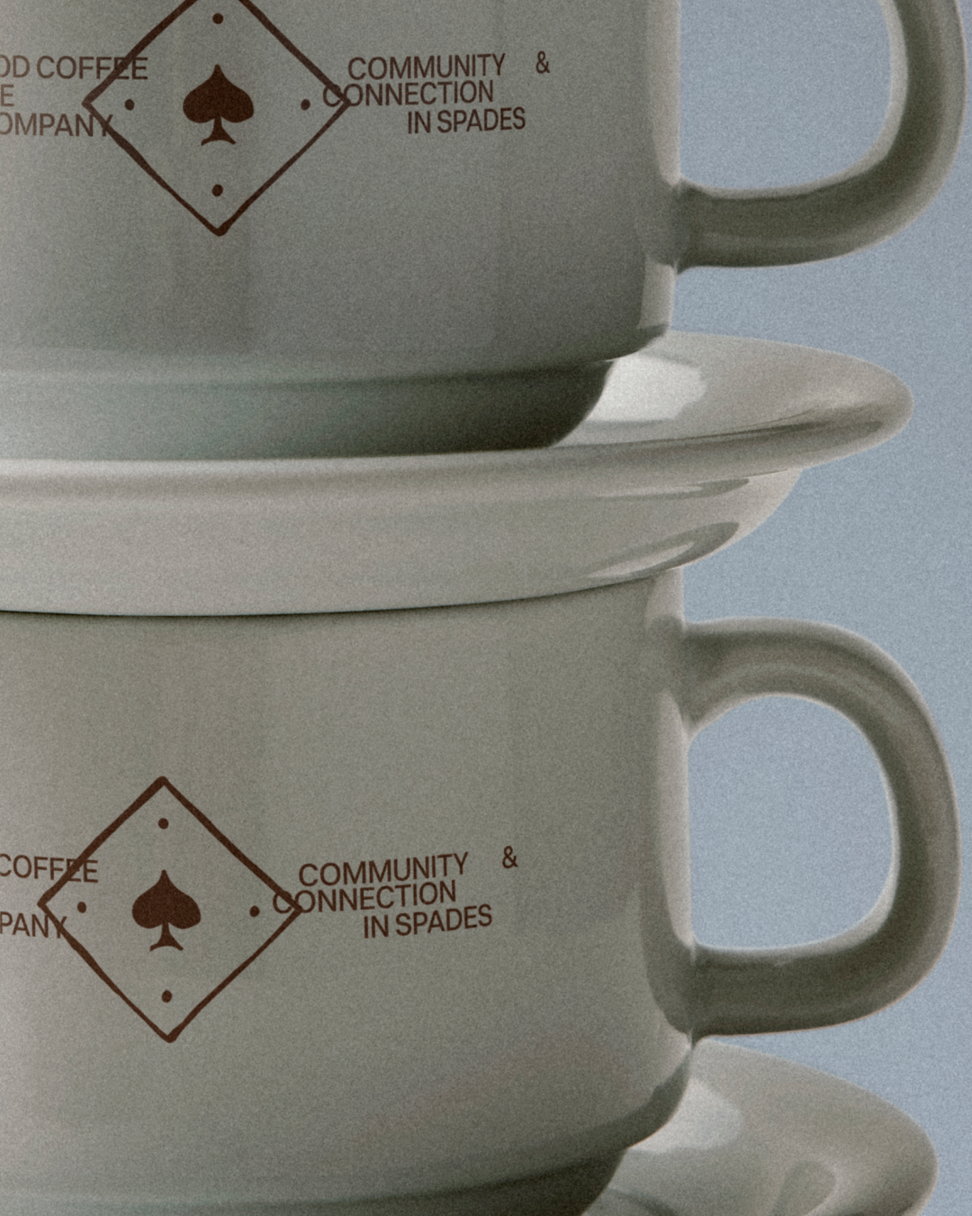 Stack of two white coffee cups with a logo and text that reads 'Community & Connection in Spades' in brown, placed on matching white saucers against a light blue background.