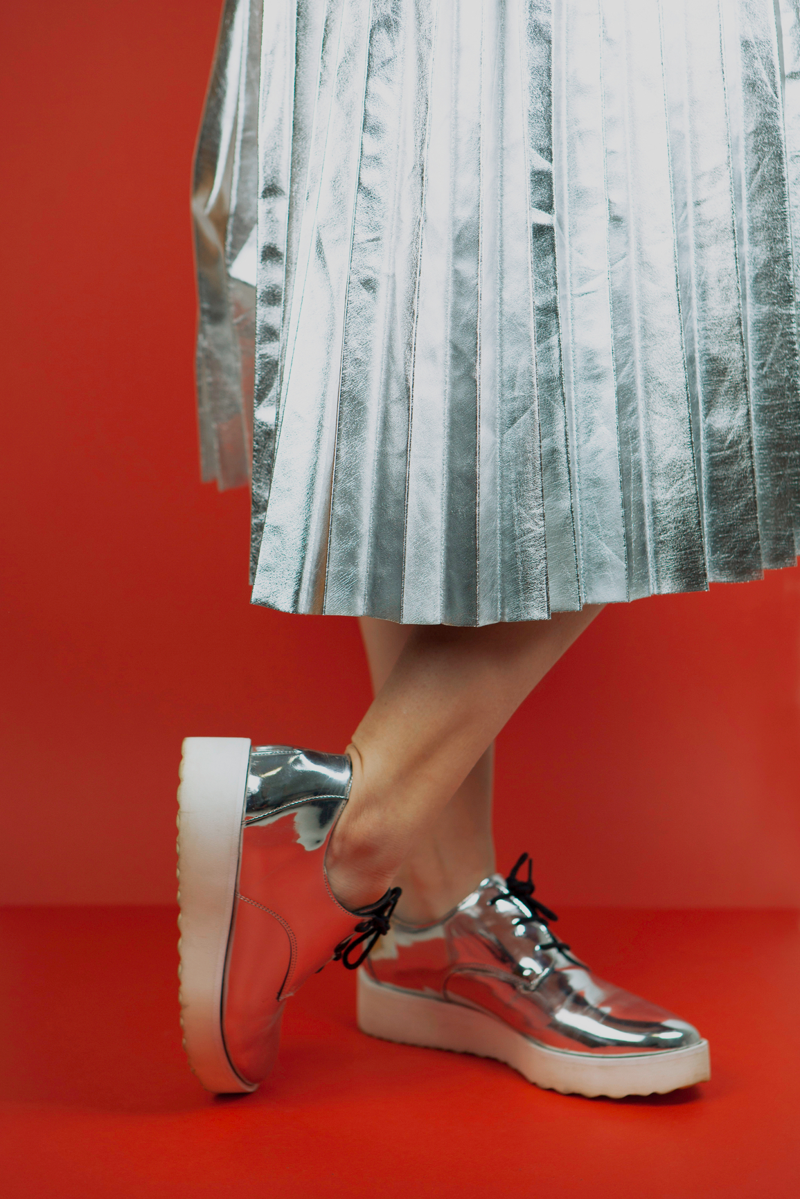 Close-up of a person's legs wearing shiny silver shoes and a silver pleated skirt, standing against a red background.