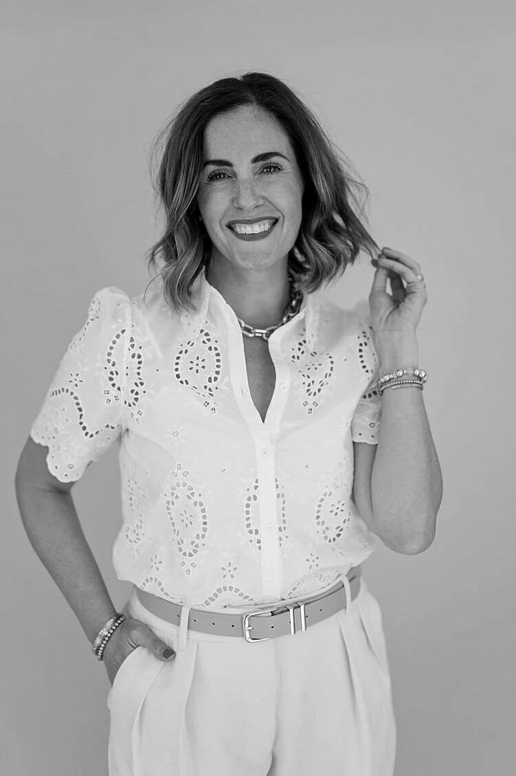 Black-and-white portrait of a woman smiling, wearing a white embroidered blouse, white pants, and jewelry, against a plain background.
