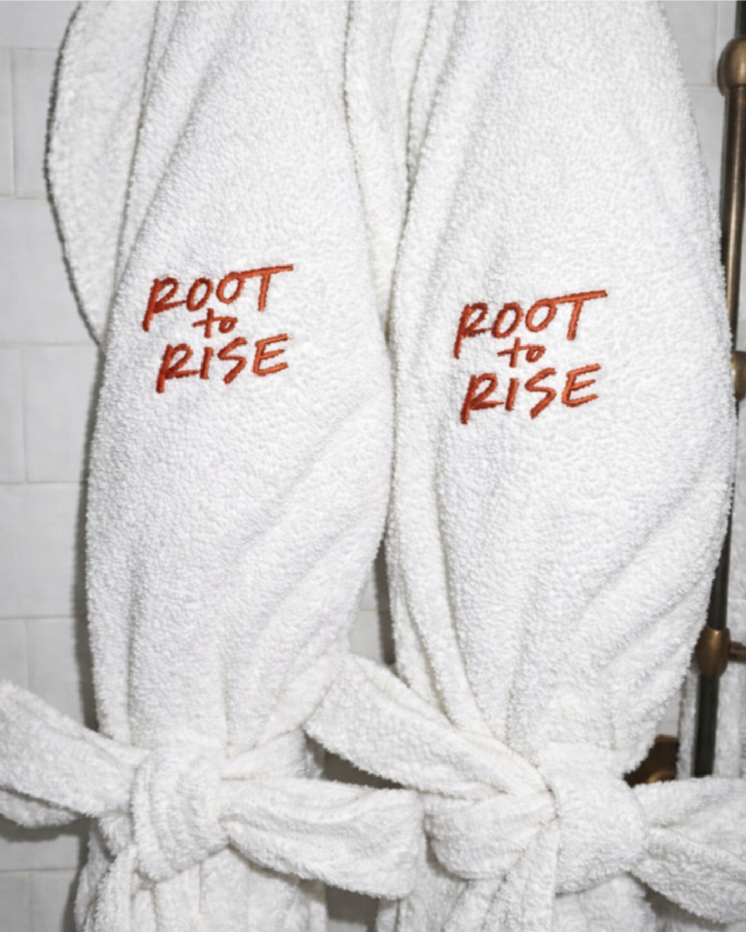 White bathrobes with red embroidered logos for 'Root to Rise', tied at the waist, hanging on a wall.
