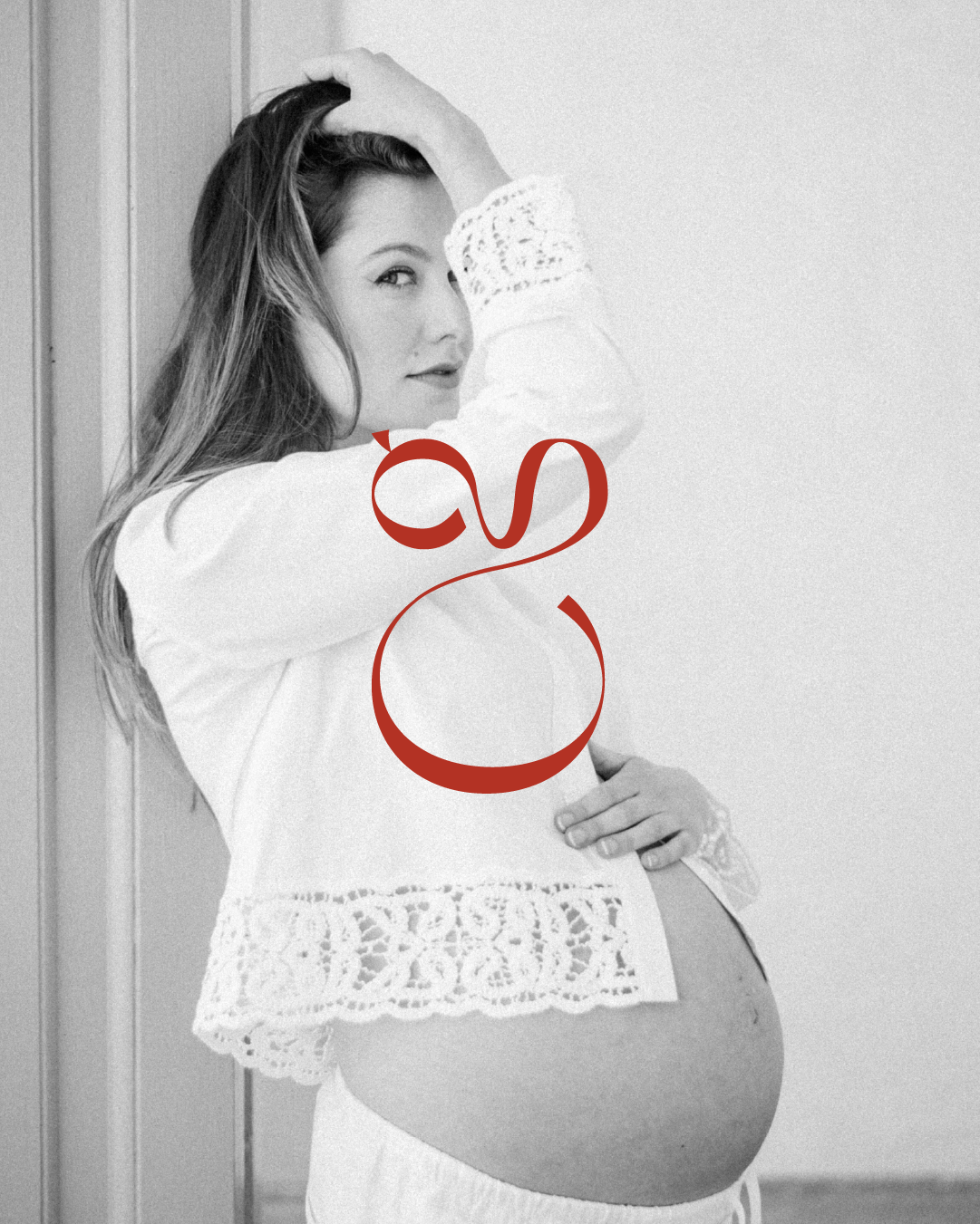 Black and white photo of a pregnant woman standing indoors with her hand on her head, wearing a white lace top and smiling and the CG logo overlayed in red.