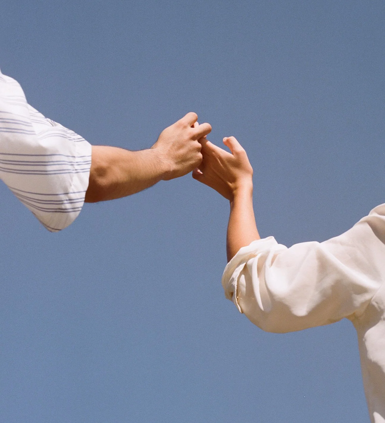 Two people holding hands reaching towards each other against a clear blue sky.