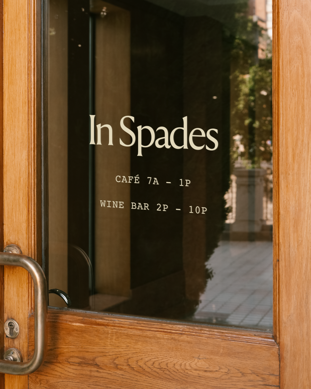 Wooden restaurant door with a glass panel displaying the logo for 'In Spades' and operating hours for a cafe and wine bar.