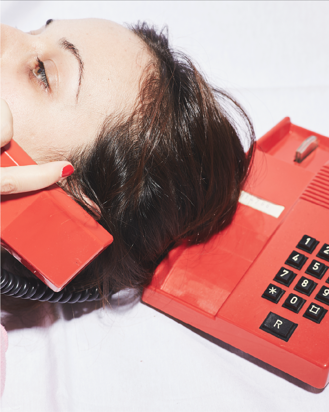 A woman with brown hair lying down, with her head resting on a red rotary phone's cradle, talking on the phone with the receiver held to her ear.