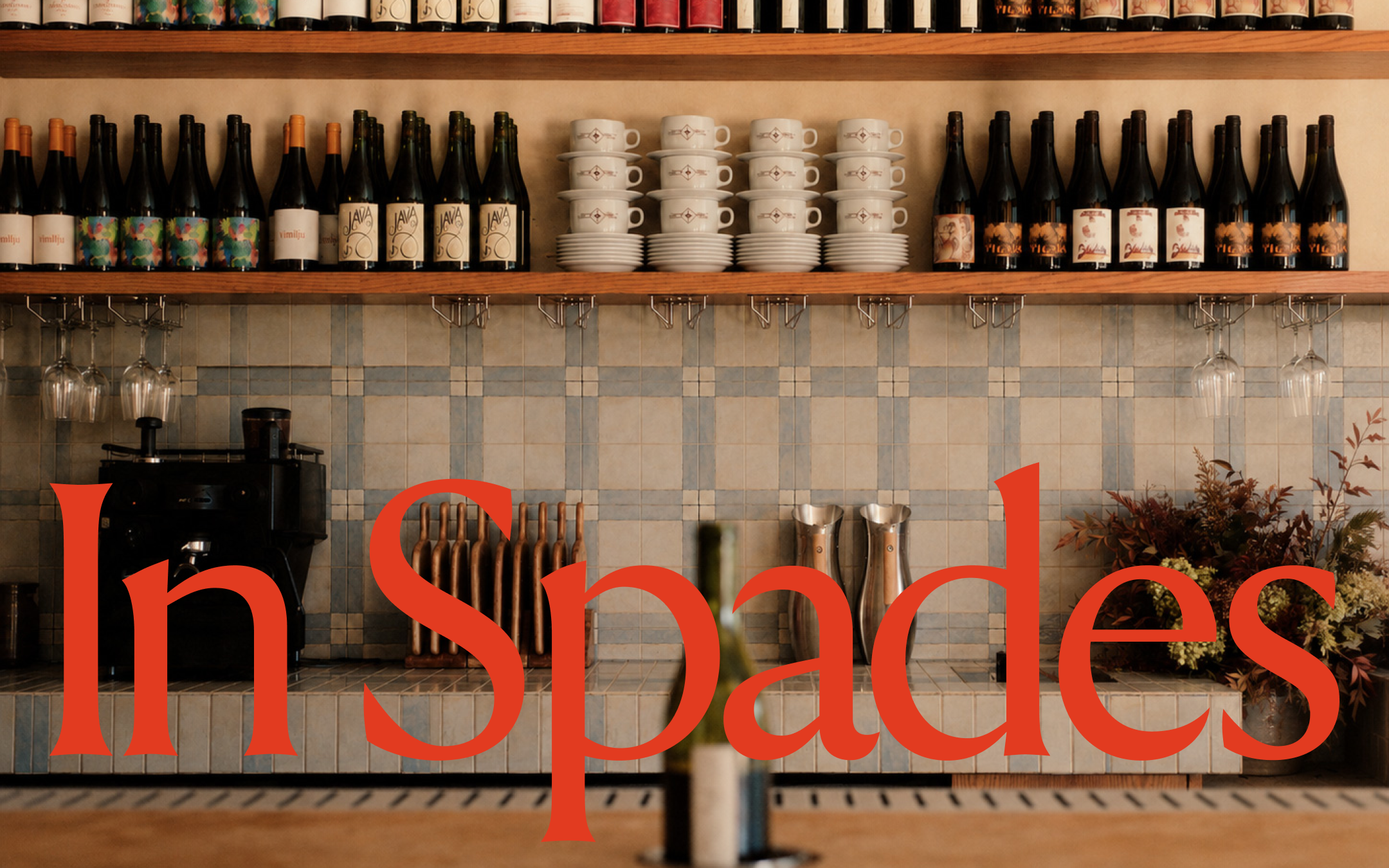 Shelves with wine bottles and stacked coffee cups in a cafe with the In Spades café and wine bar logo overlayed in red on top