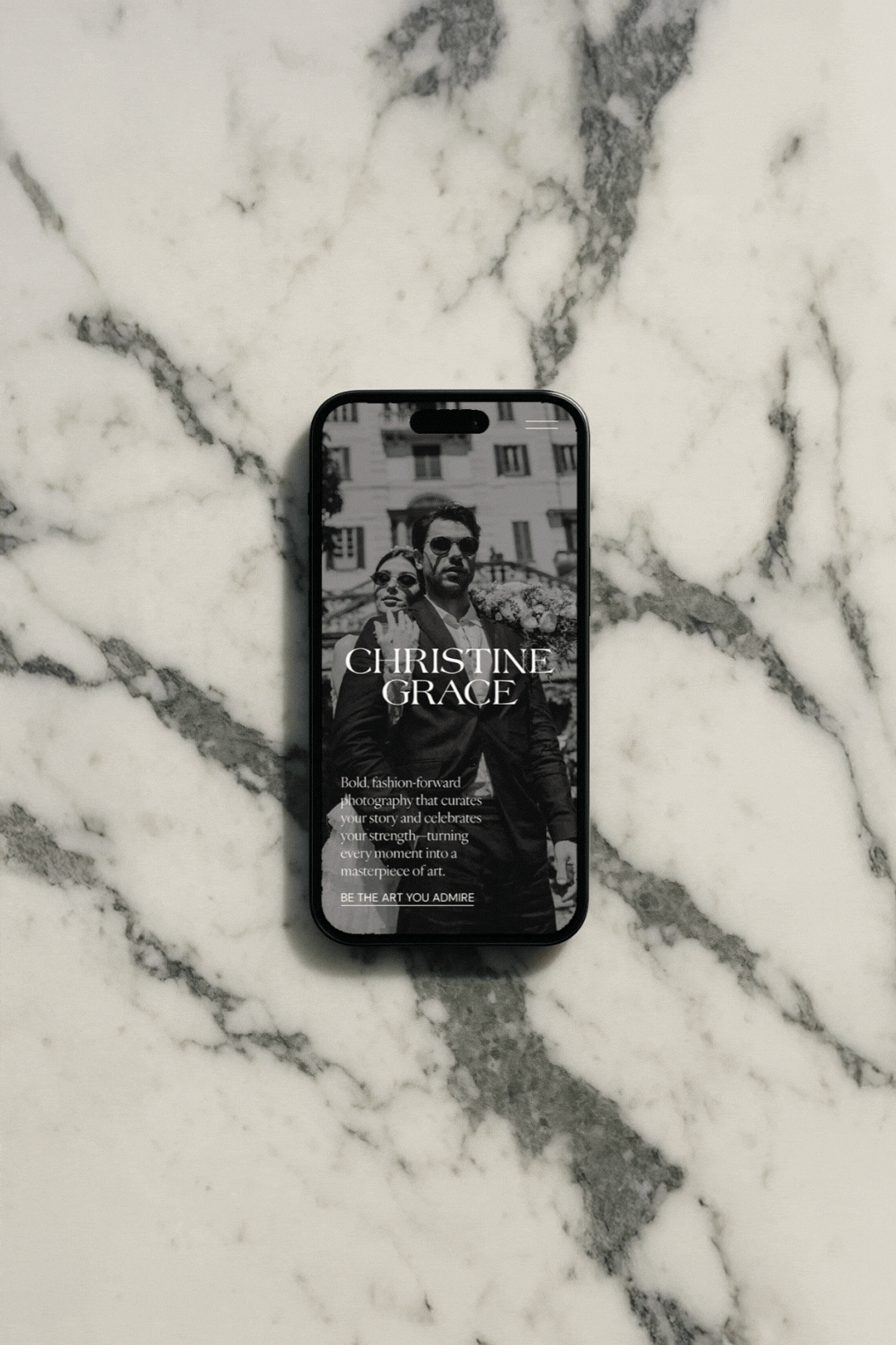 A smartphone displaying a black-and-white fashion photography website with a woman and man in stylish outfits, set against a city background, on a white marble surface.