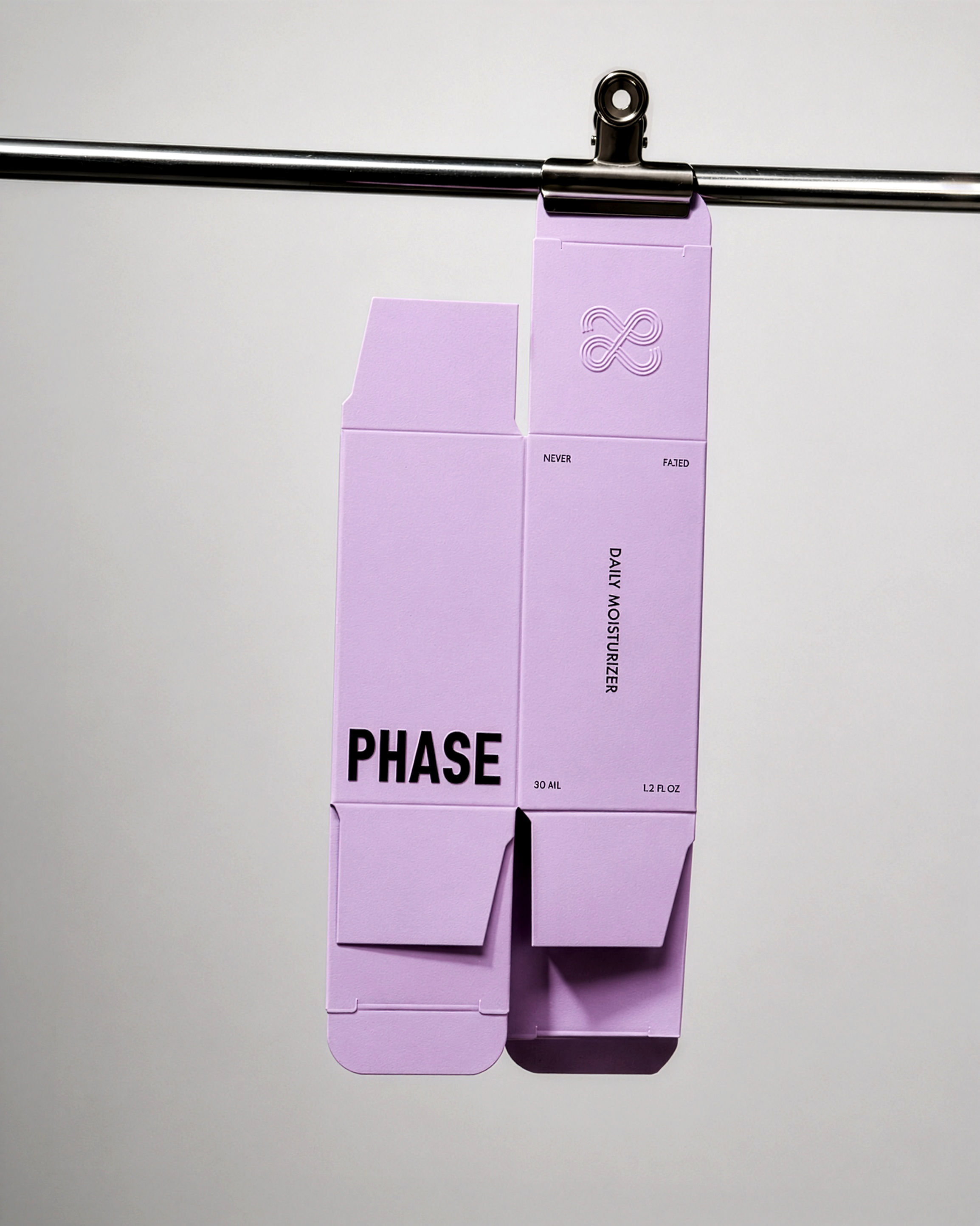 A lilac skincare packaging box with black Phase logo and 'DAILY MOISTURIZER' attached to a metal rod with a black clip.