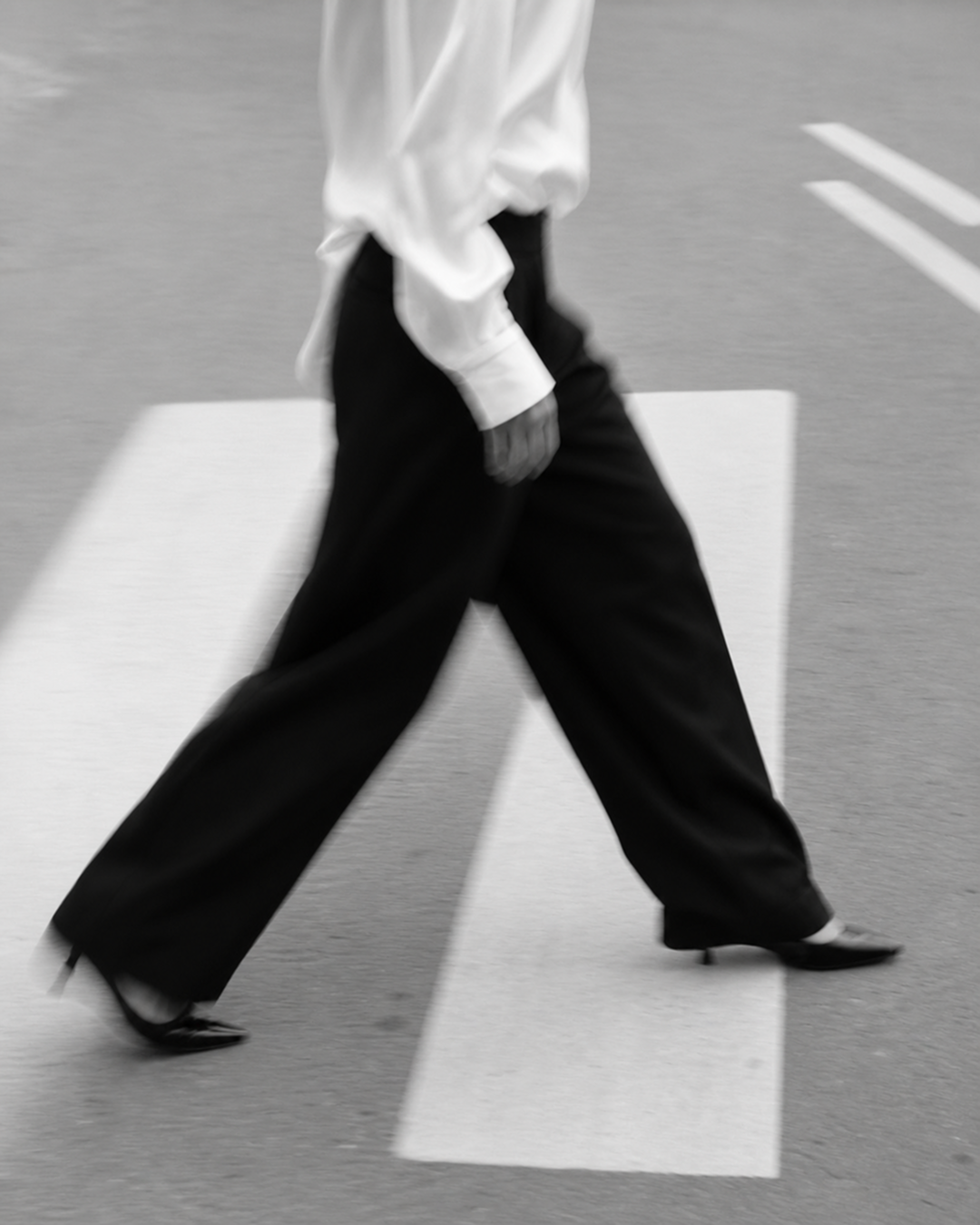 Person walking on crosswalk in black and white, wearing a white shirt and black trouser pants.