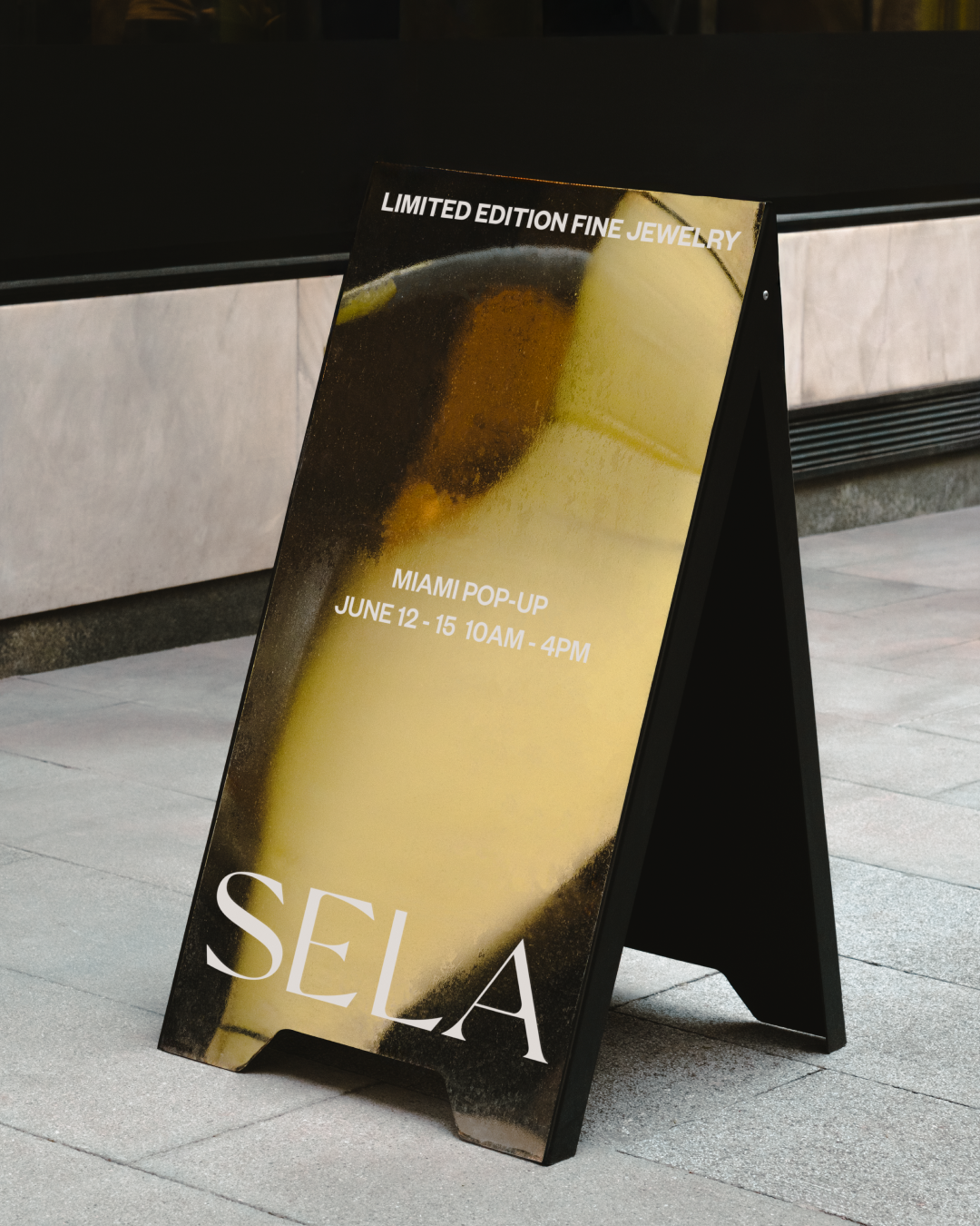 A black A-frame signboard with a poster advertising a limited edition fine jewelry event in Miami, occurring from June 12 to 15, from 10 AM to 4 PM. The signboard is made of mirror-like material.