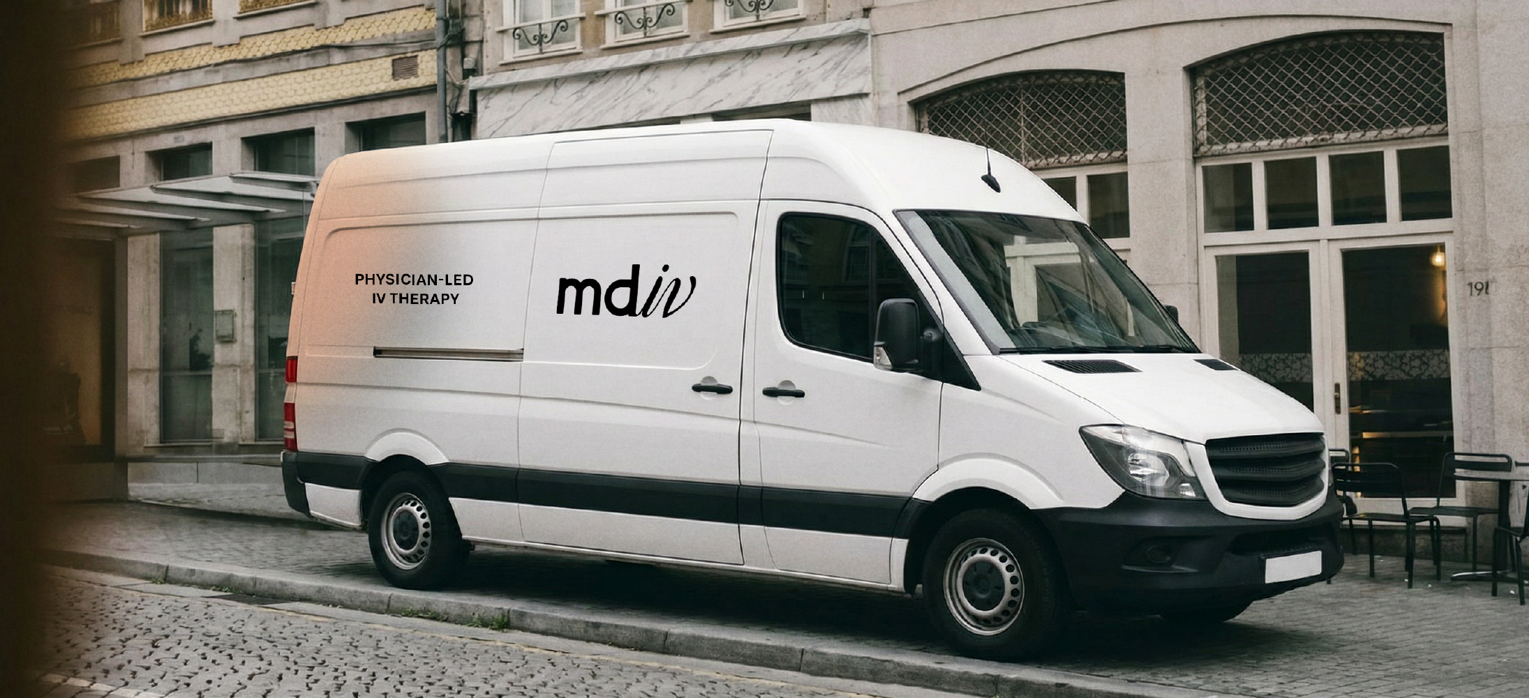 A branded white cargo van parked on a cobblestone street in front of a building with large windows. The van has black text on its side that reads 'PHYSICIAN-LED IV THERAPY' and a stylized logo that reads 'mdiv'.