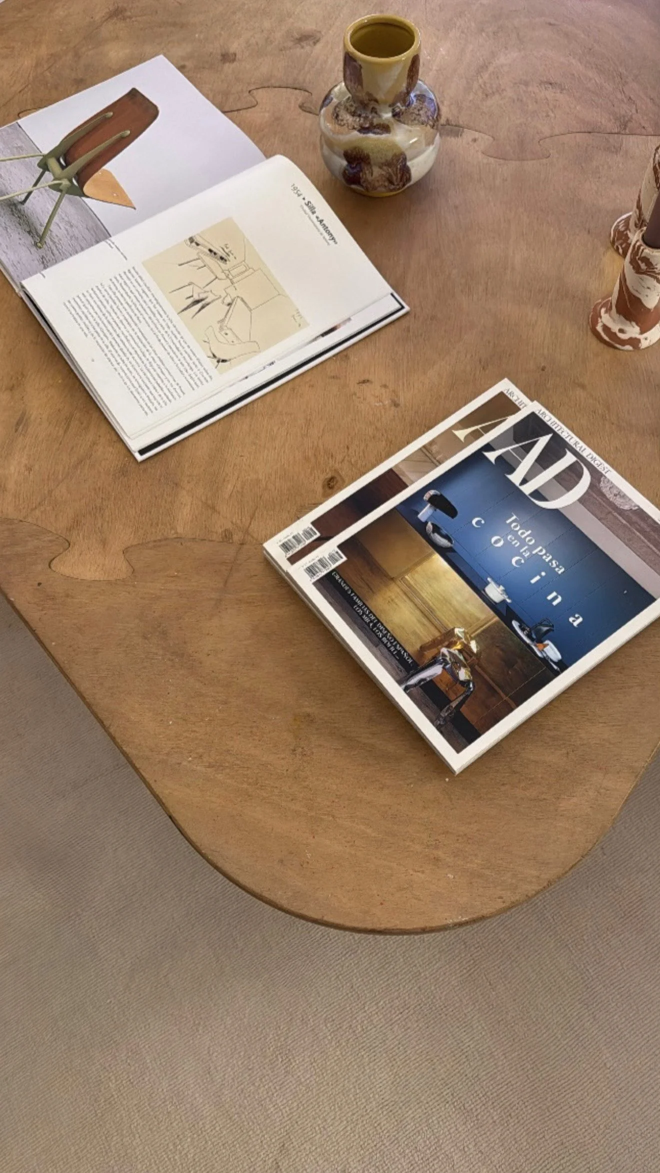 A wooden table with a magazine titled 'AD' related to interior design, an open book with illustrations and text, placed beside a ceramic vase with a marbled pattern, and a small decorative ceramic cup.