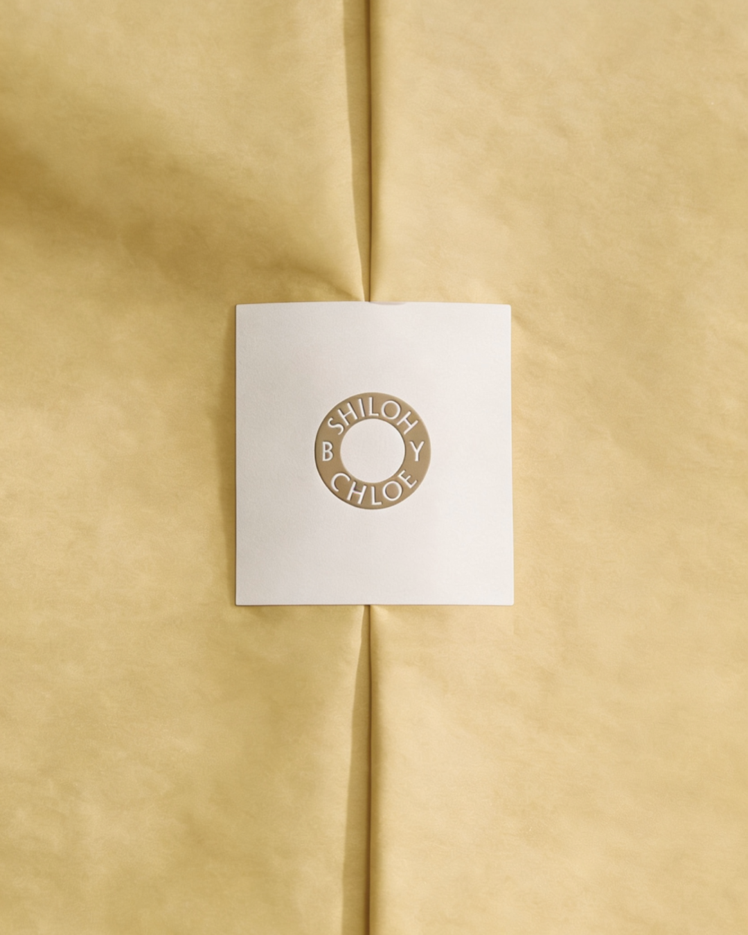 A white sticker with a circular gold brand mark featuring the words "SHILOH BY CHLOE" placed on yellow tissue paper