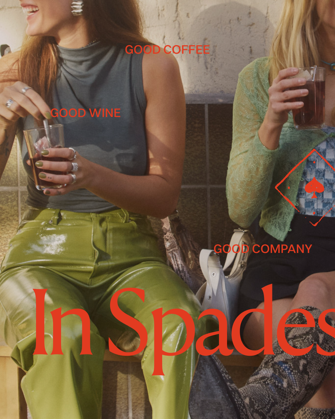 Two women sitting together, holding drinks. One woman has a grey sleeveless top and shiny green pants, smiling. The other woman is wearing a green lace top and holding a glass of dark beverage. There are labels on the image: "Good Wine" "Good Coffee", and "Good Company". In Spades is overlaid at the bottom of the image.