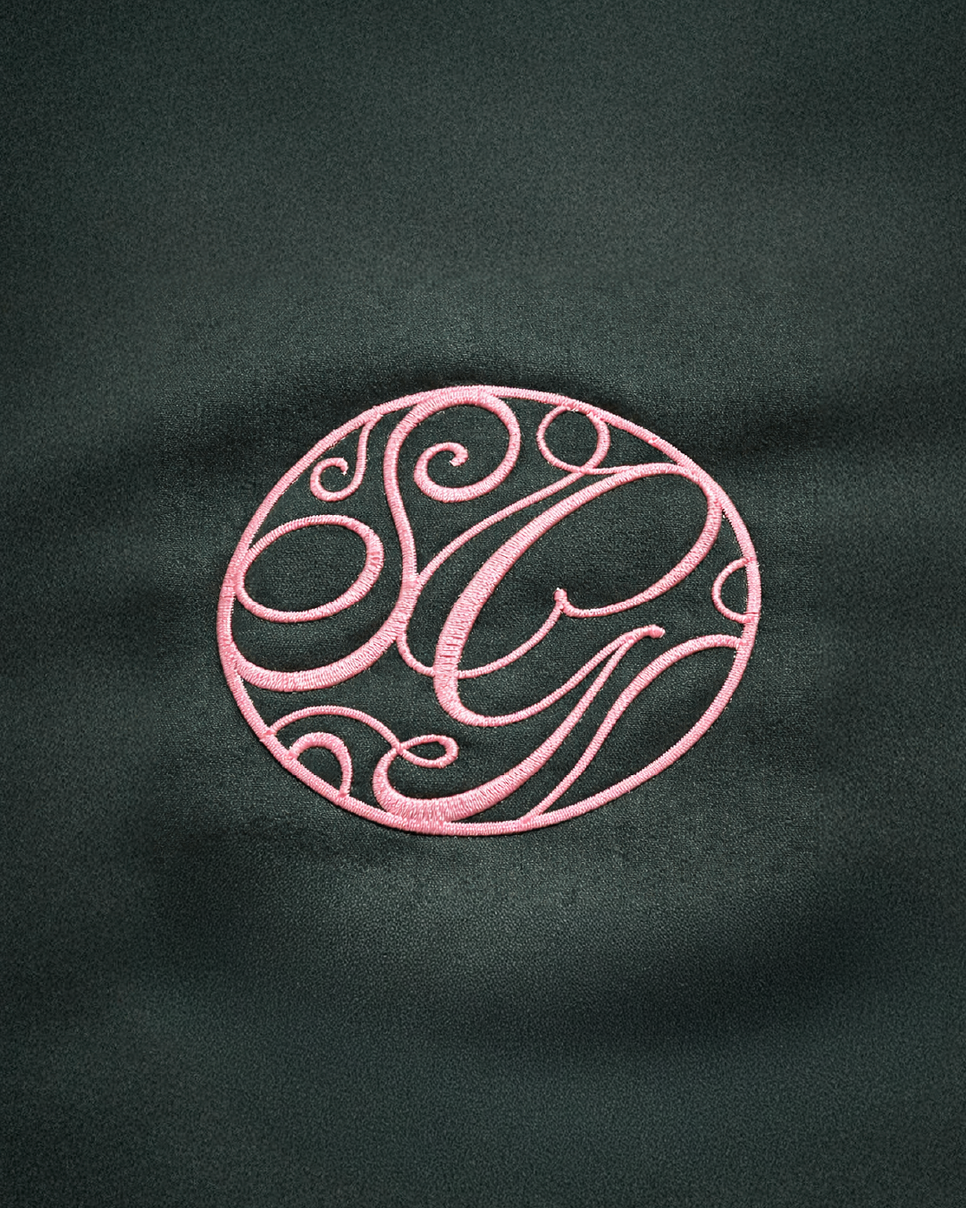 Pink embroidered monogram with initials inside a circle on green fabric background.