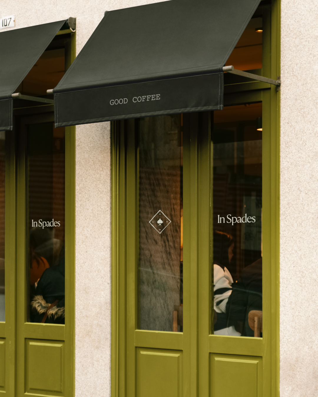 Outside of a coffee shop with green-framed windows and a black awning that reads 'Good Coffee'; inside, customers sit at tables visible through the glass.