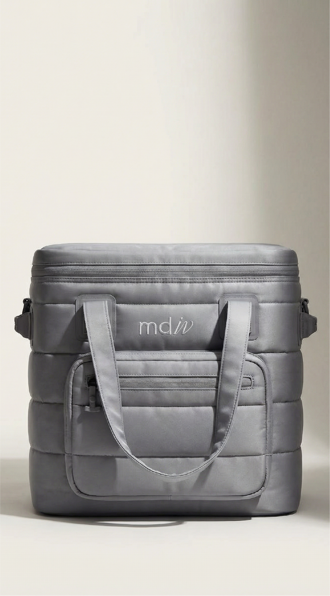 A branded gray quilted cooler bag with handles and a front zip pocket, sitting on a white surface. The bag has a silver logo patch with the logo for MD IV iv therapy and wellness brand