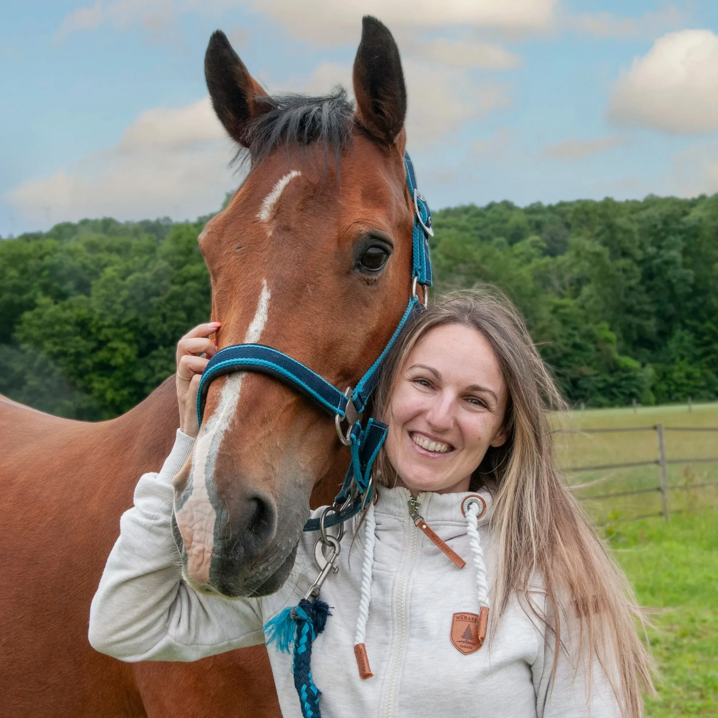 A woman with long hair smiling, standing next to a brown horse with a gray mane, in a field with green trees and a cloudy sky.