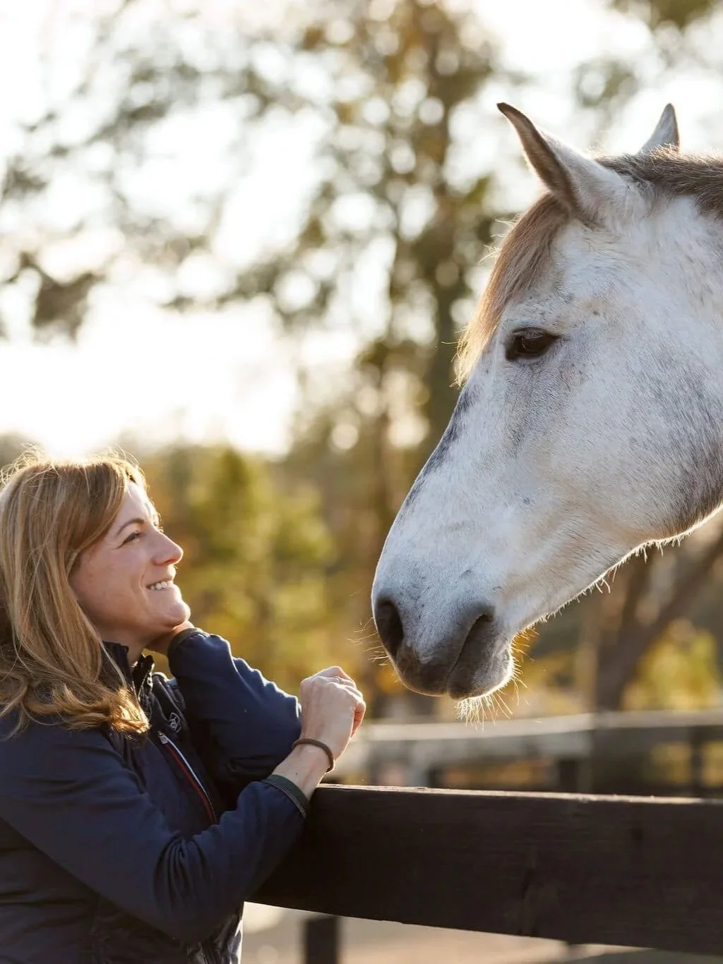 A woman smiling and interacting with a white horse at a farm or nature setting during sunset.