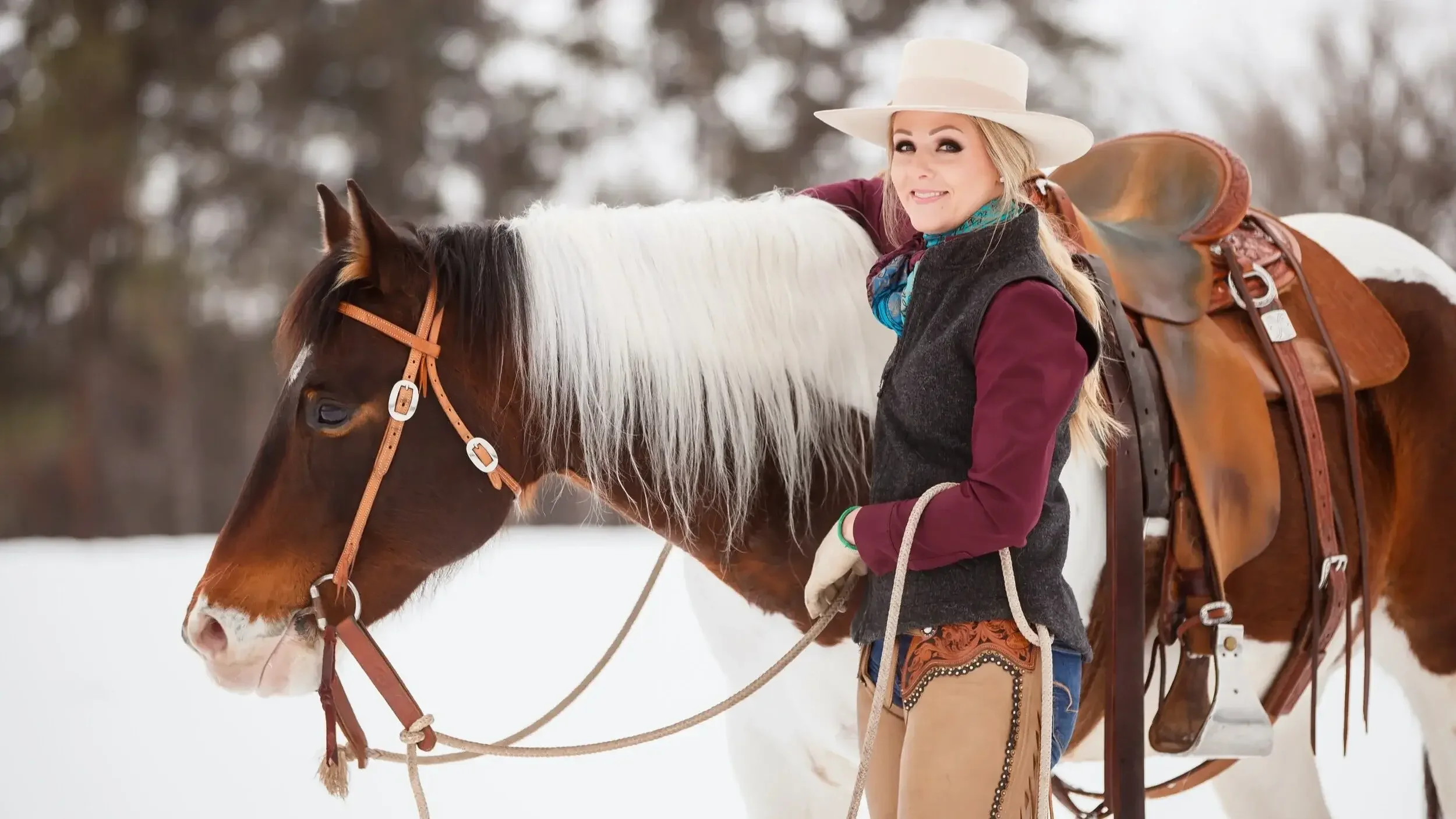 A woman with blonde hair wearing a white cowboy hat, a blue scarf, and western attire standing next to a brown and white horse in a snowy outdoor setting with blurred trees in the background.