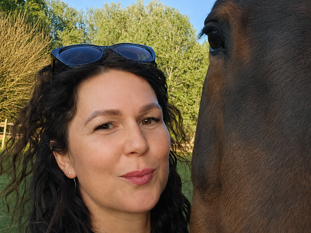 A woman with dark curly hair, sunglasses on her head, and earrings taking a selfie with a brown horse outdoors during daytime. The background features green trees and a clear blue sky.